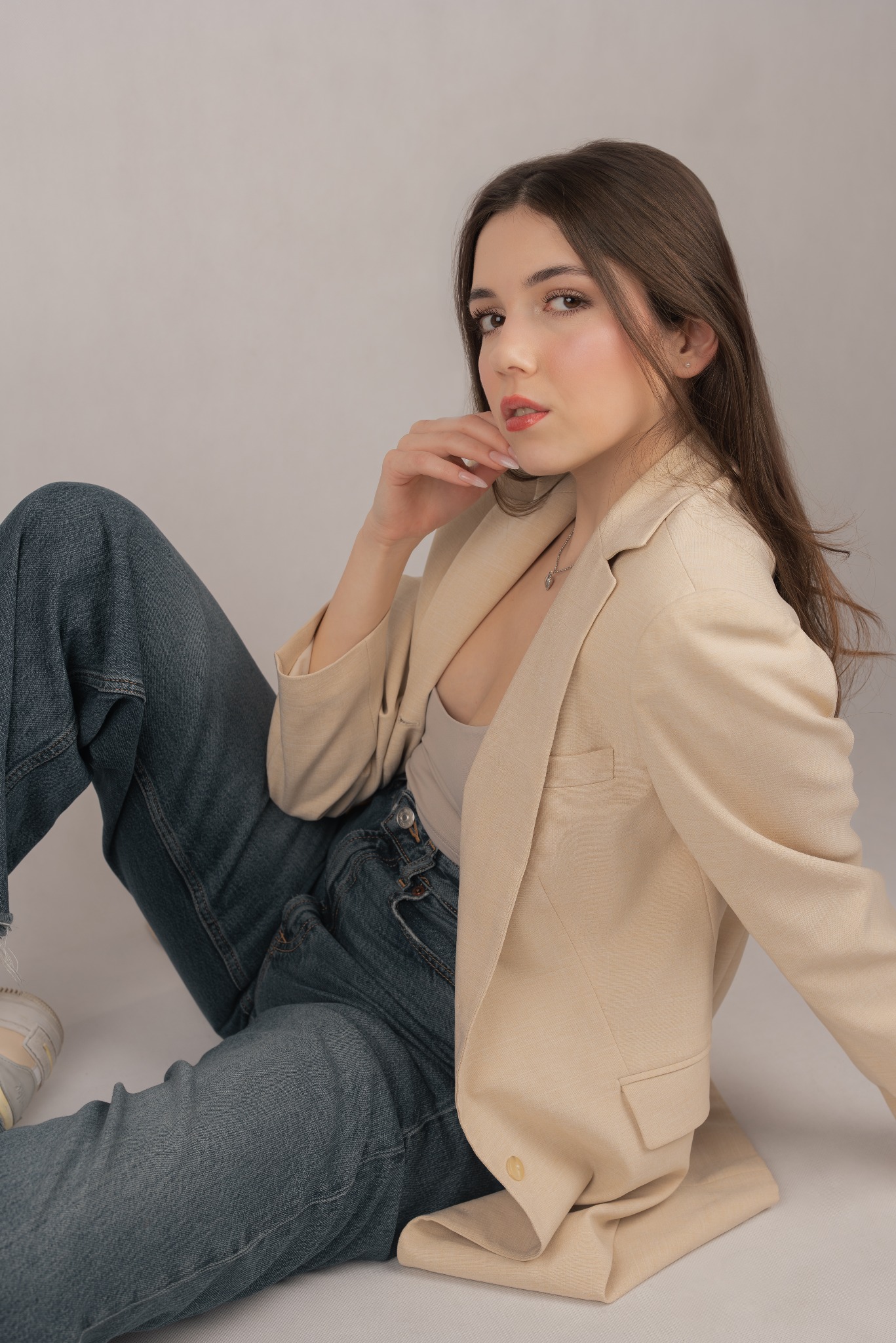 Portret kobiety w beżowej marynarce i jeansach, siedzącej na podłodze. Ujęcie z dołu, podkreślające spojrzenie i naturalny makijaż. Stonowana kolorystyka.
