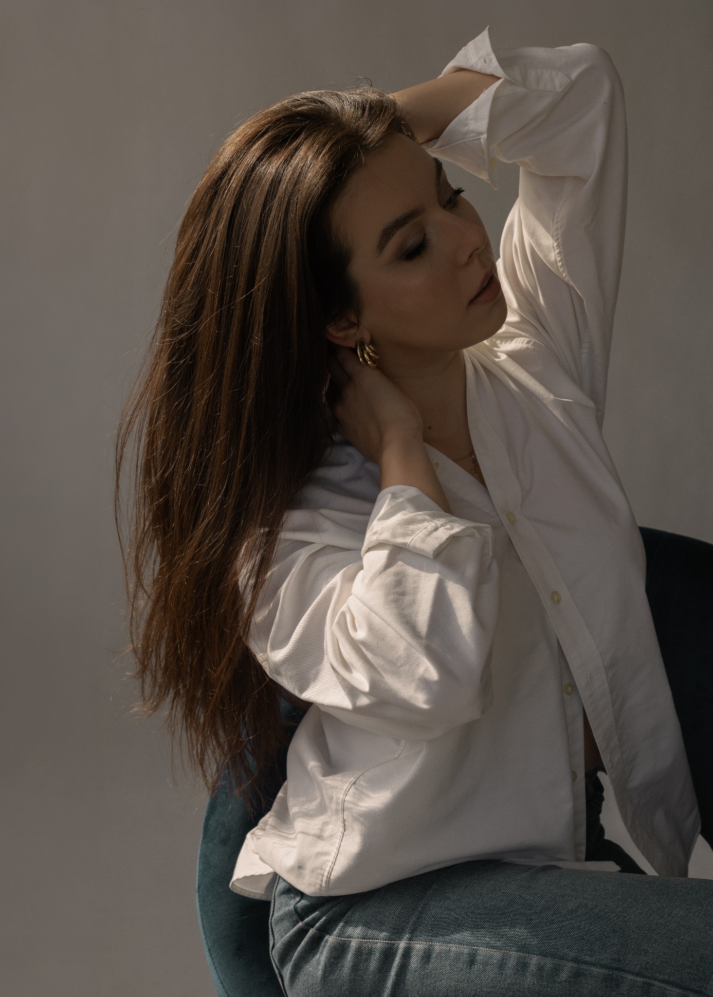 Portret kobiety w białej koszuli i jeansach, siedzącej na krześle. Delikatne światło podkreśla jej rysy twarzy i złote kolczyki. Ujęcie z profilu, z rękami uniesionymi do góry.