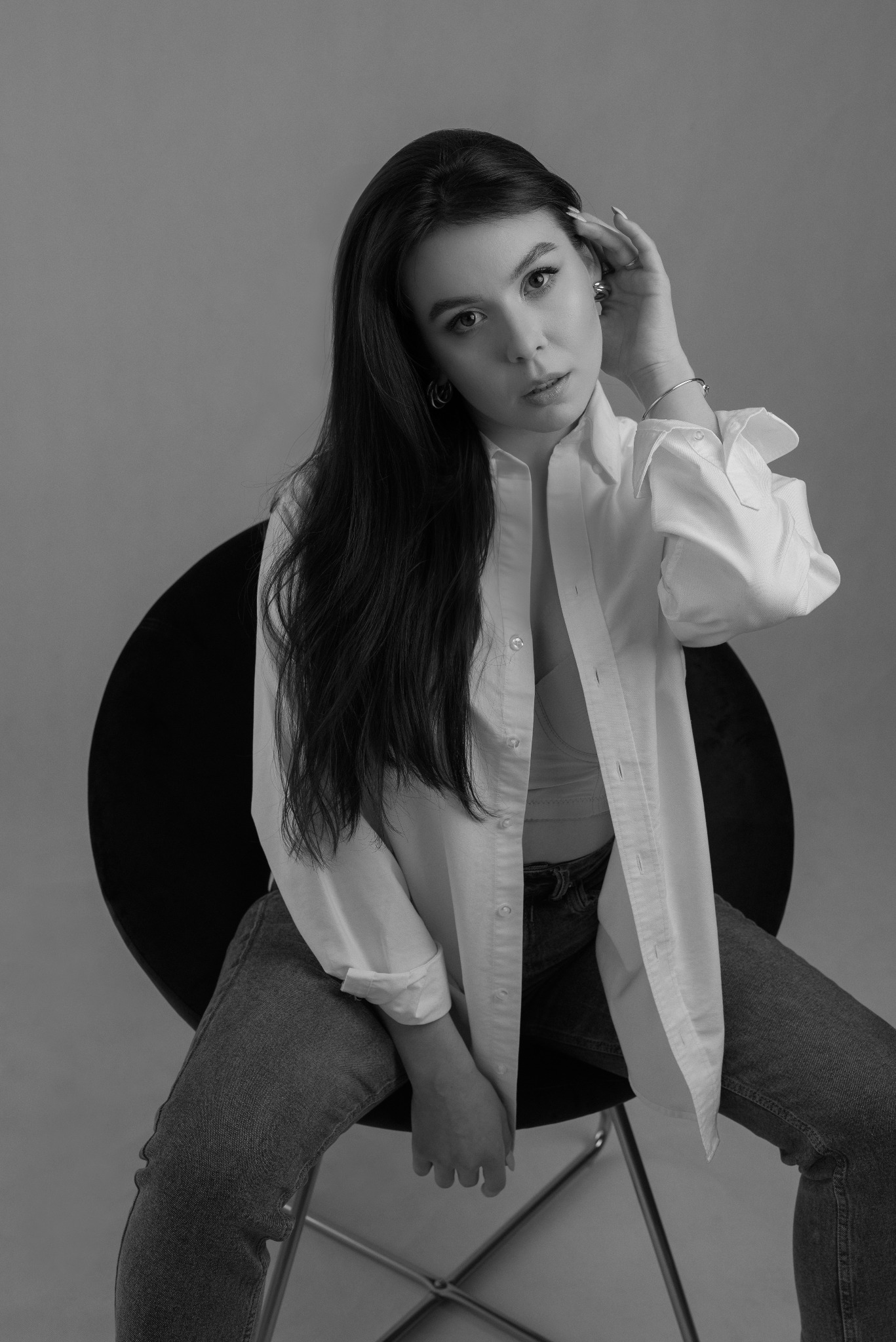 Monochromatyczny portret kobiety siedzącej na krześle, ubrana w rozpiętą białą koszulę i jeansy, z ręką uniesioną do włosów, patrząca w obiektyw.
