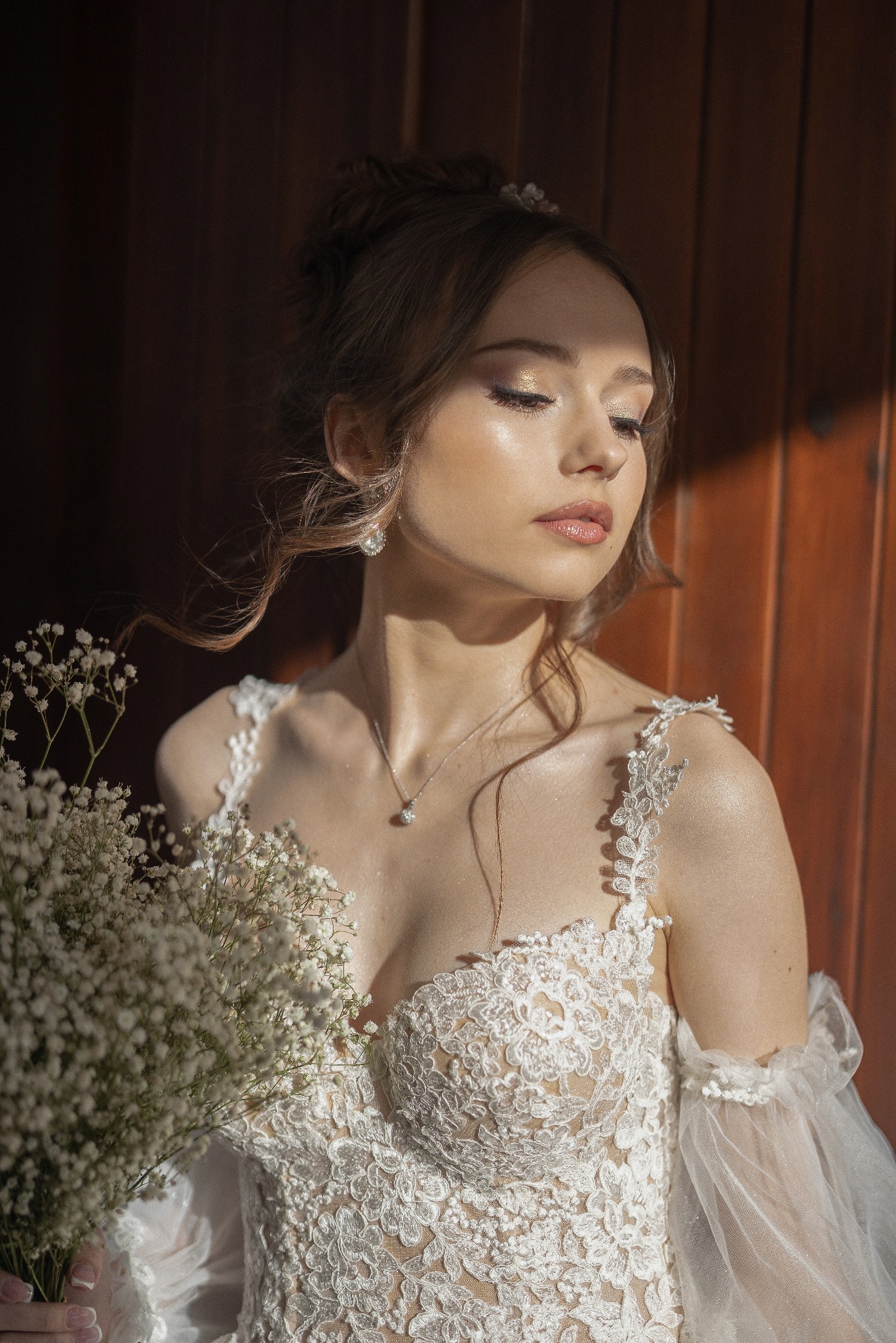 Panna młoda w koronkowej sukni, z bukietem gipsówki, w ciepłym świetle na tle drewnianej ściany. Subtelny makijaż i delikatna biżuteria podkreślają uroczysty charakter.