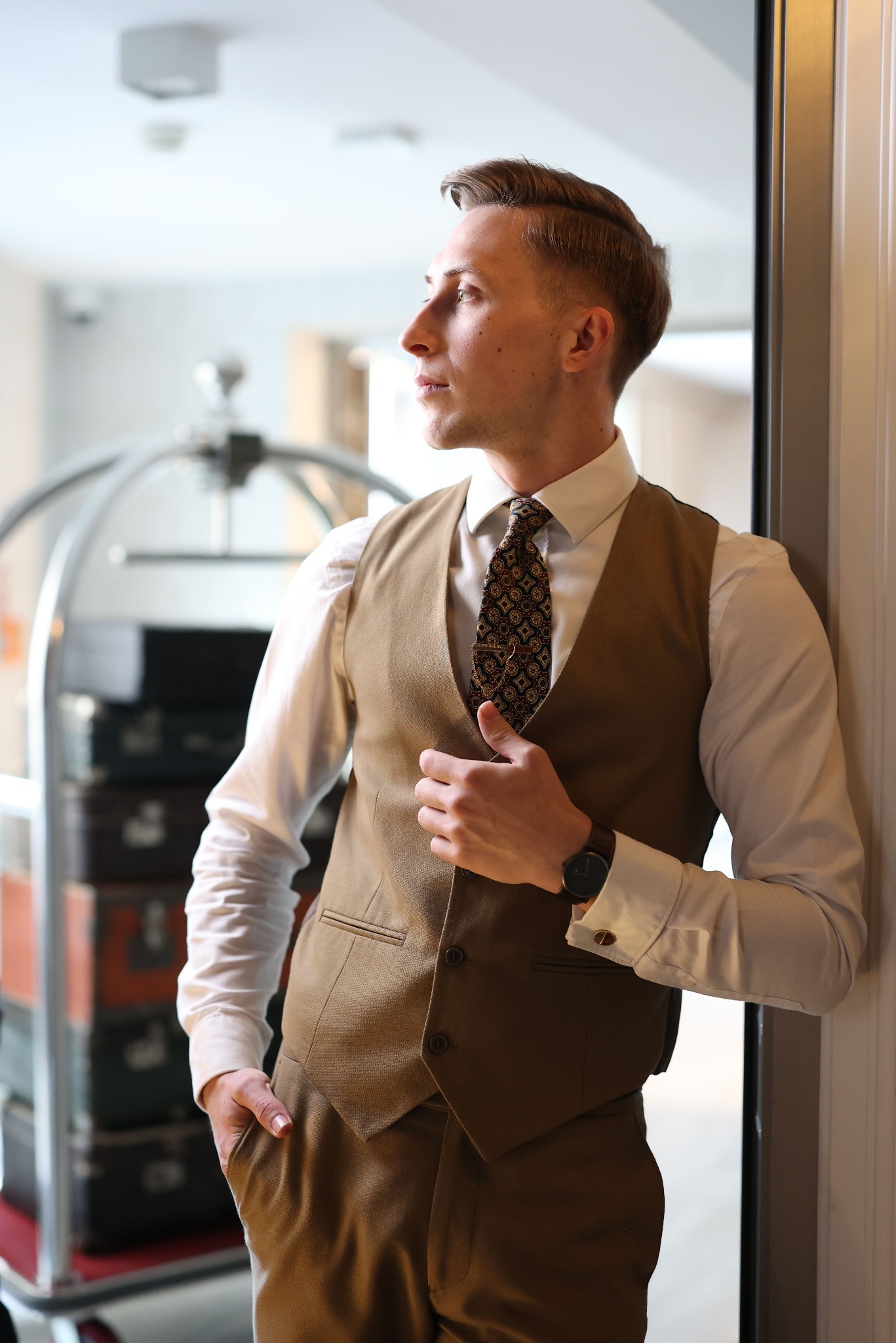 Mężczyzna w brązowym garniturze i krawacie, oparty o ścianę, z wózkiem bagażowym w tle. Elegancki strój, stonowana kolorystyka, naturalne światło.