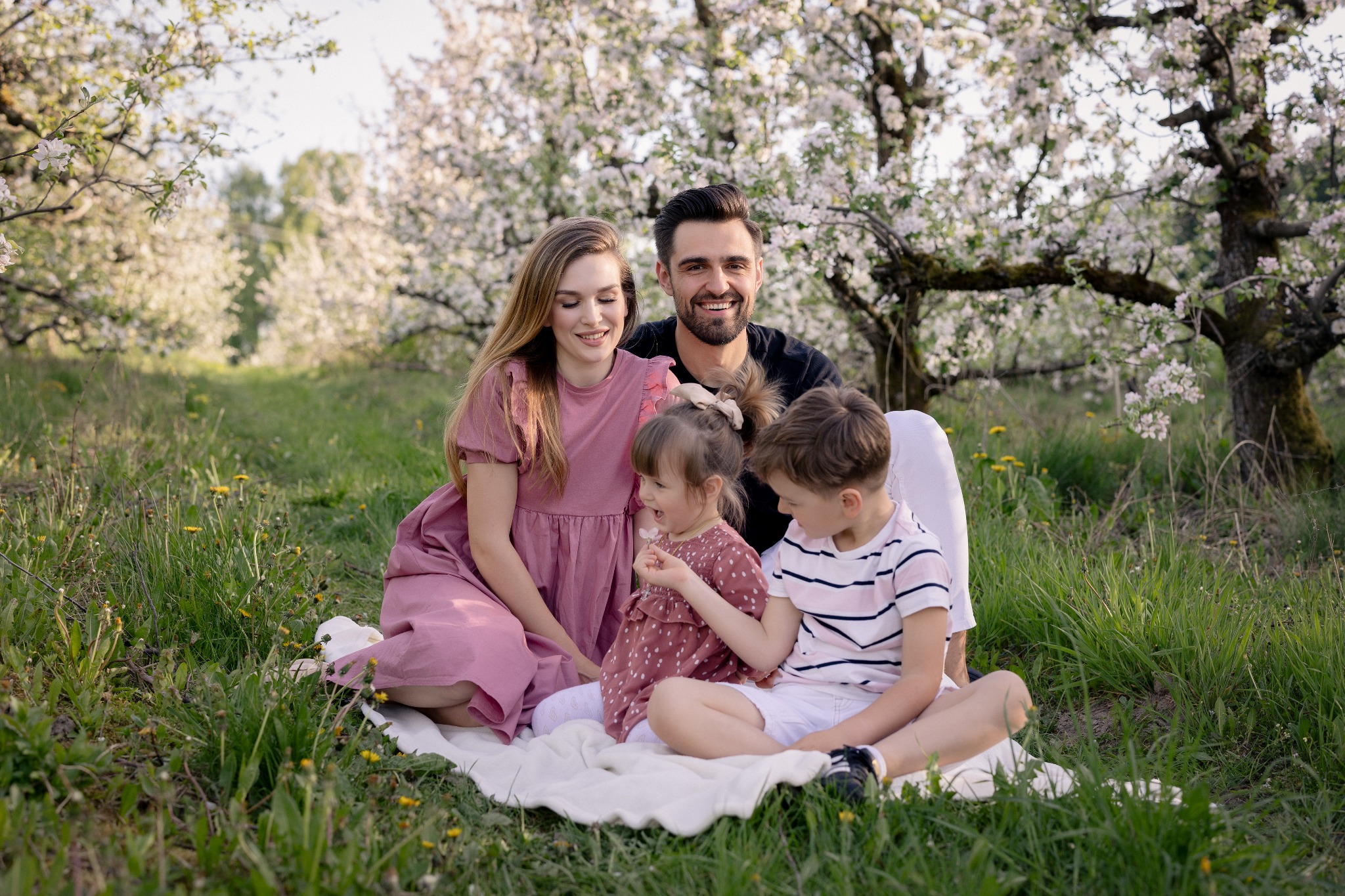 Uśmiechnięta rodzina (mama, tata, córka i syn) siedzi na kocu w sadzie kwitnących drzew. Mama w sukience w kolorze dusty rose, tata w czarnej koszulce, dzieci w pasujących strojach.