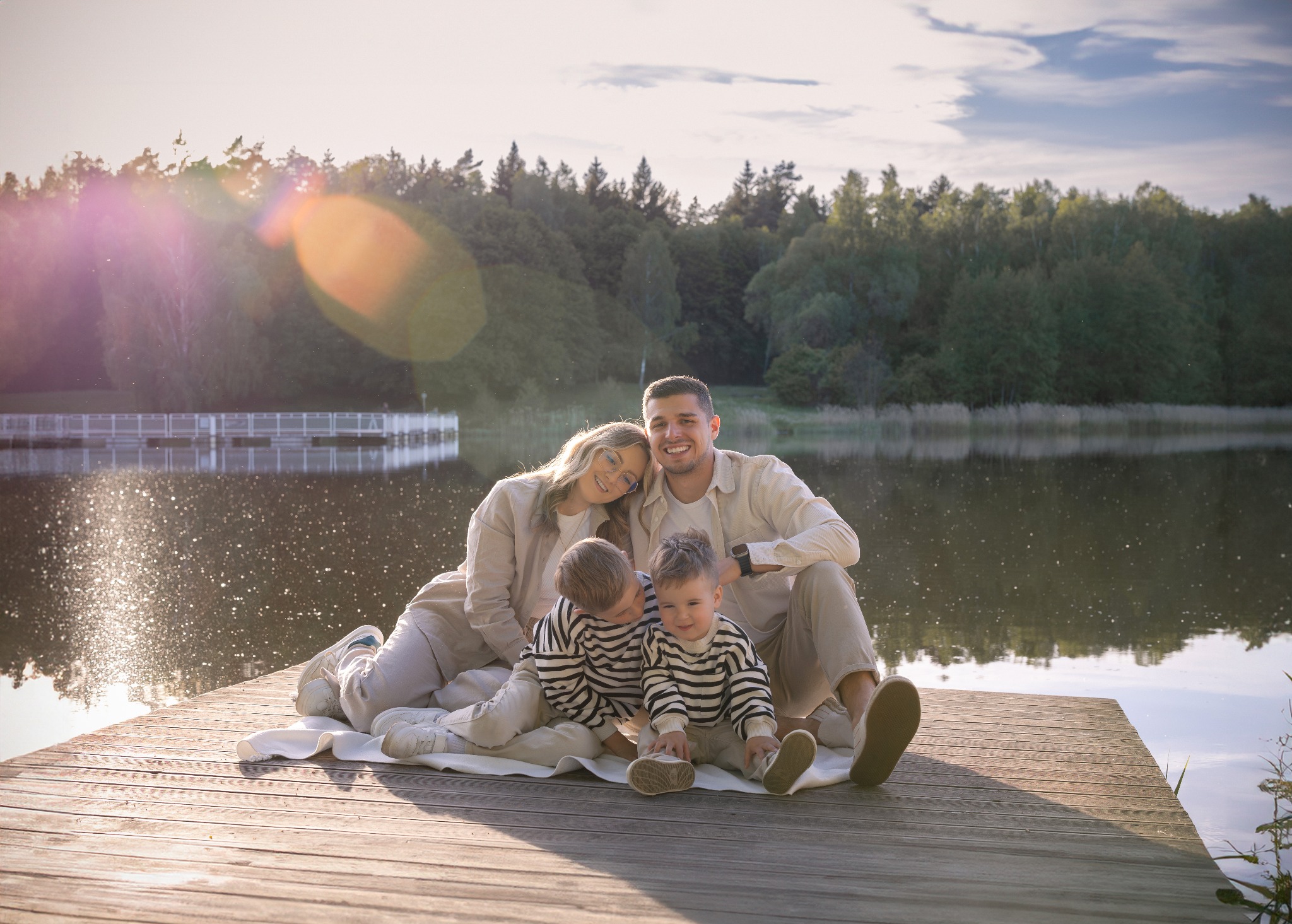 Uśmiechnięta rodzina z dwójką dzieci siedzi na drewnianym pomoście nad jeziorem w ciepłym, naturalnym świetle. W tle widoczny las i błękitne niebo z chmurami.