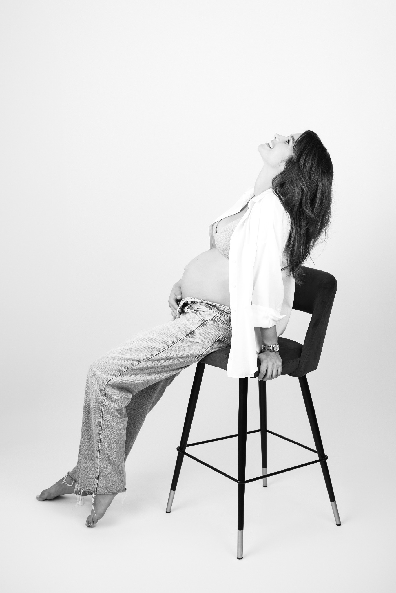 Czarno-białe zdjęcie kobiety w ciąży, siedzącej na krześle barowym, z głową odchyloną do tyłu i uśmiechem na twarzy. Ubrana w jeansy i białą koszulę, z ręką na brzuchu.