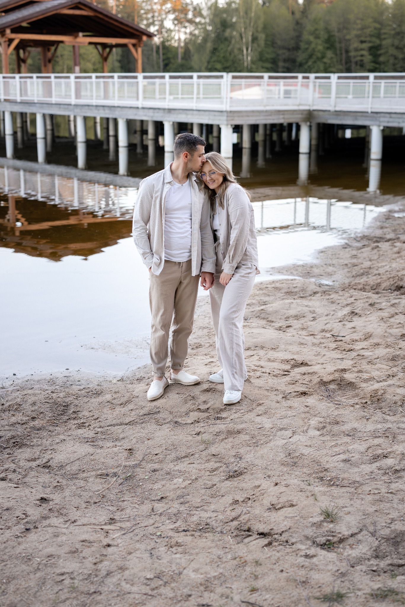 Zakochana para w beżowych ubraniach na piaszczystej plaży, mężczyzna całuje kobietę w czoło, w tle drewniany pomost nad wodą. Romantyczna atmosfera, naturalne światło.