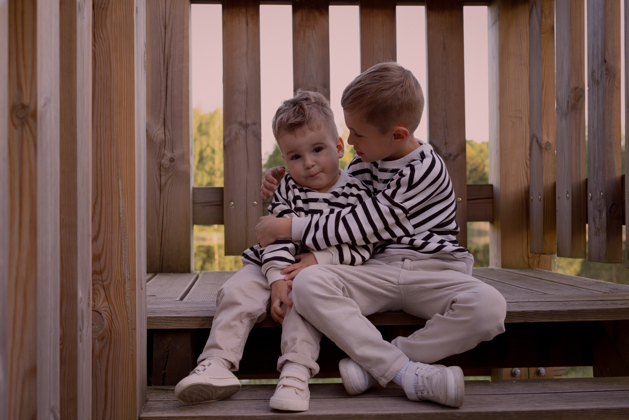 Dwóch chłopców w pasiaste bluzy siedzi na drewnianych schodach konstrukcji. Starszy obejmuje młodszego. Tło stanowią drewniane elementy i zieleń.