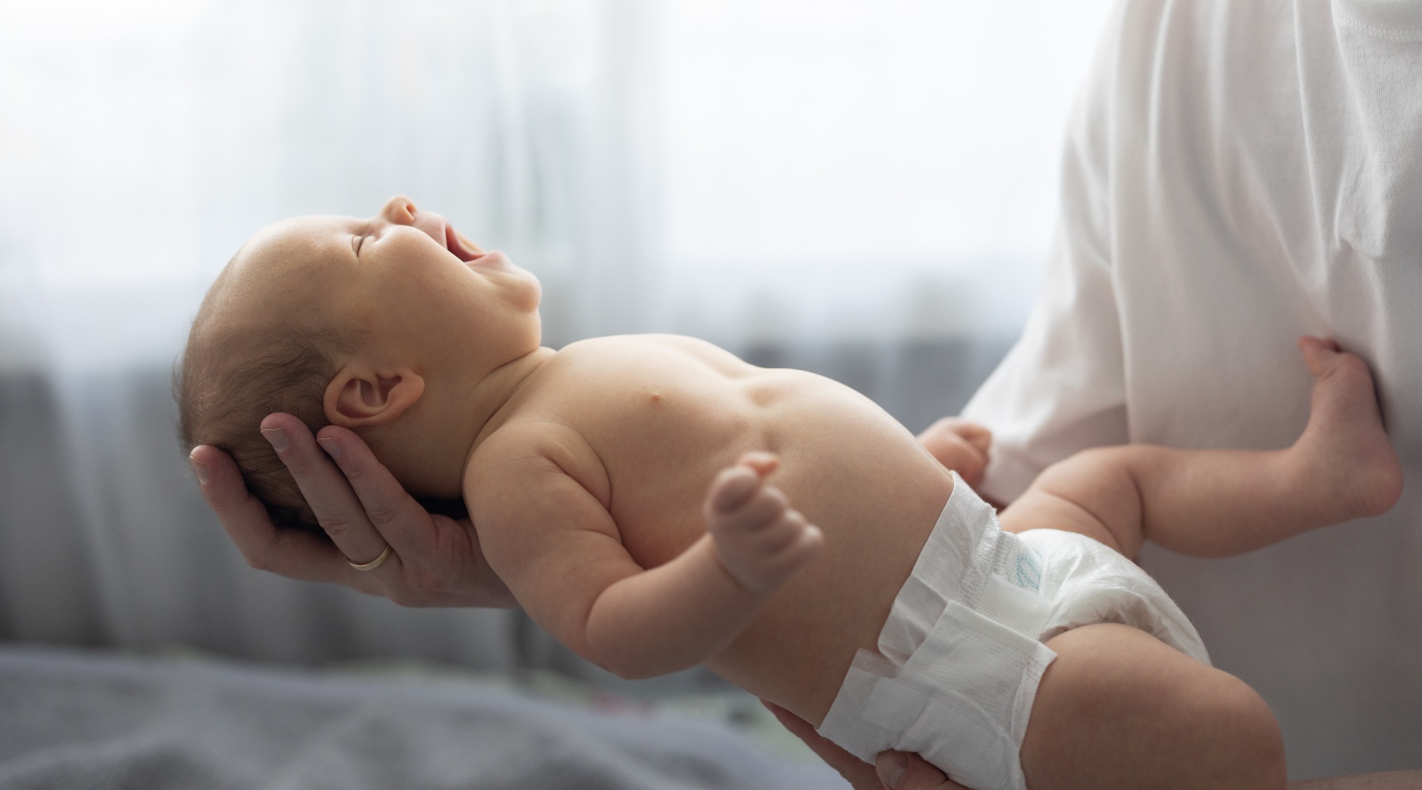 Uśmiechnięty noworodek w pieluszce, trzymany na rękach przez osobę z obrączką, w jasnym, naturalnym świetle. Dziecko z otwartymi ustami, w tle okno.