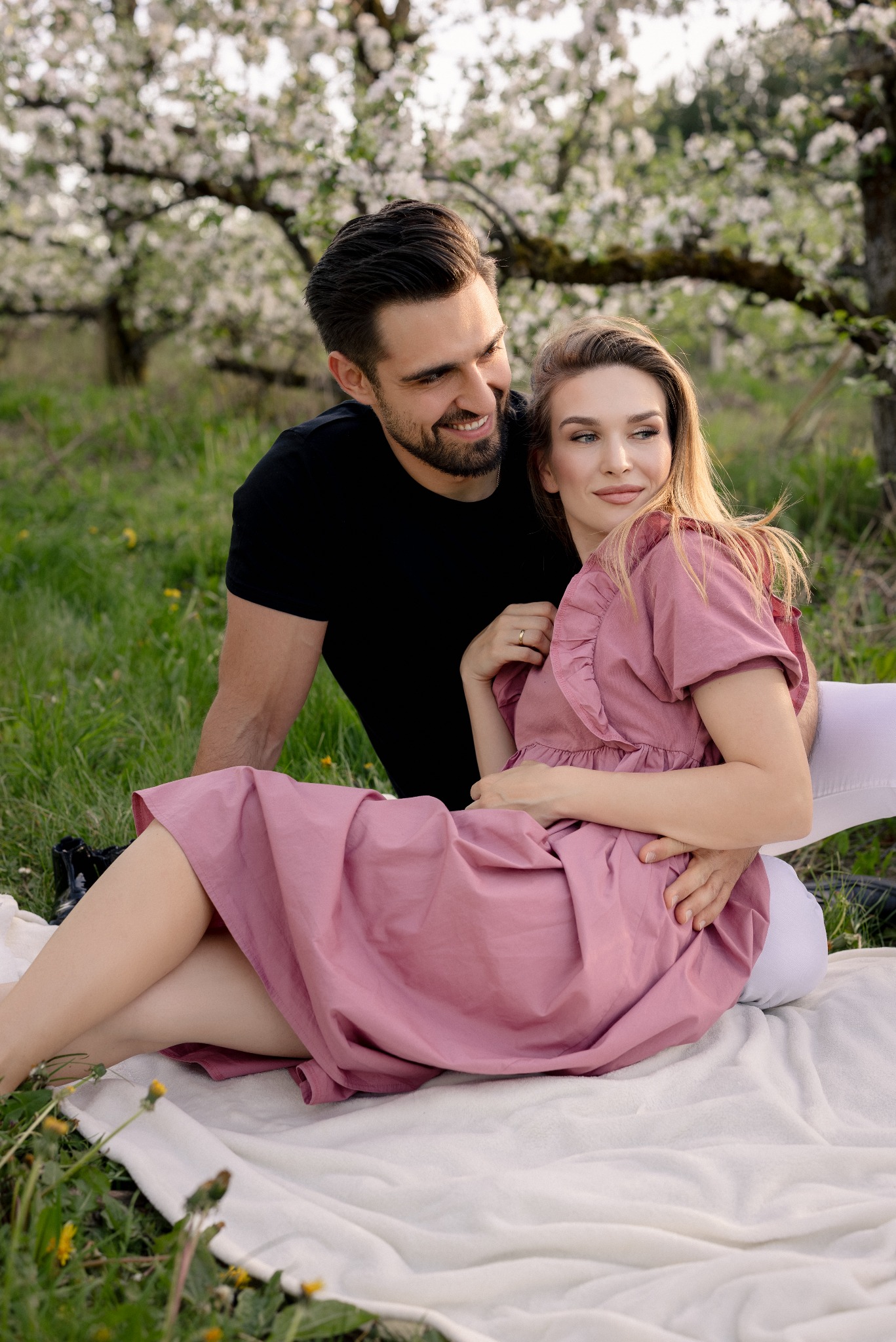 Zakochana para w sadzie jabłoni w rozkwicie. Kobieta w różowej sukience siedzi na kocu, mężczyzna obejmuje ją, oboje patrzą w dal. Romantyczna sesja w plenerze.