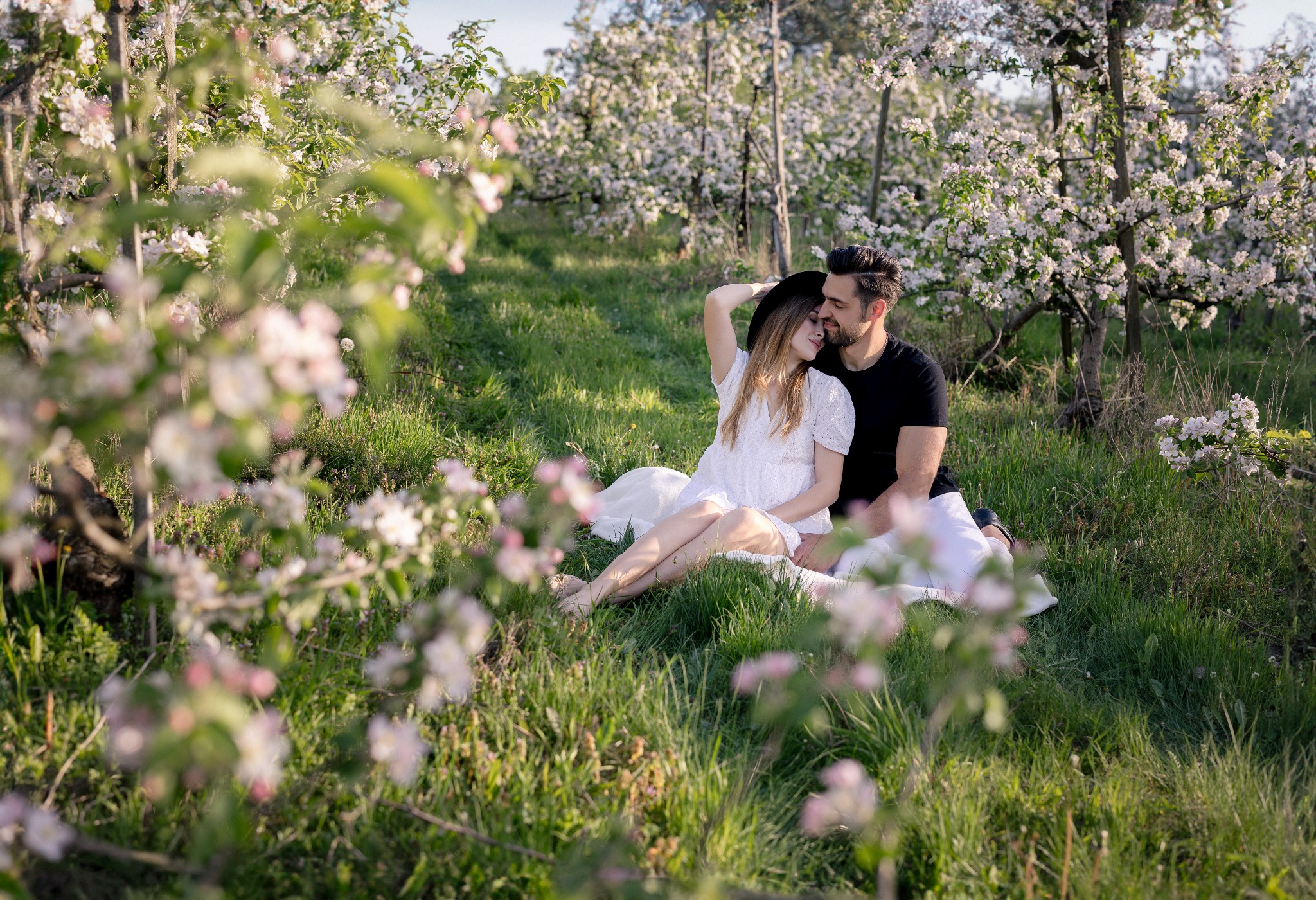 Zakochana para siedzi na trawie w sadzie kwitnących drzew. Kobieta w białej sukience i kapeluszu opiera głowę o mężczyznę w czarnej koszulce. Romantyczna sesja w plenerze.