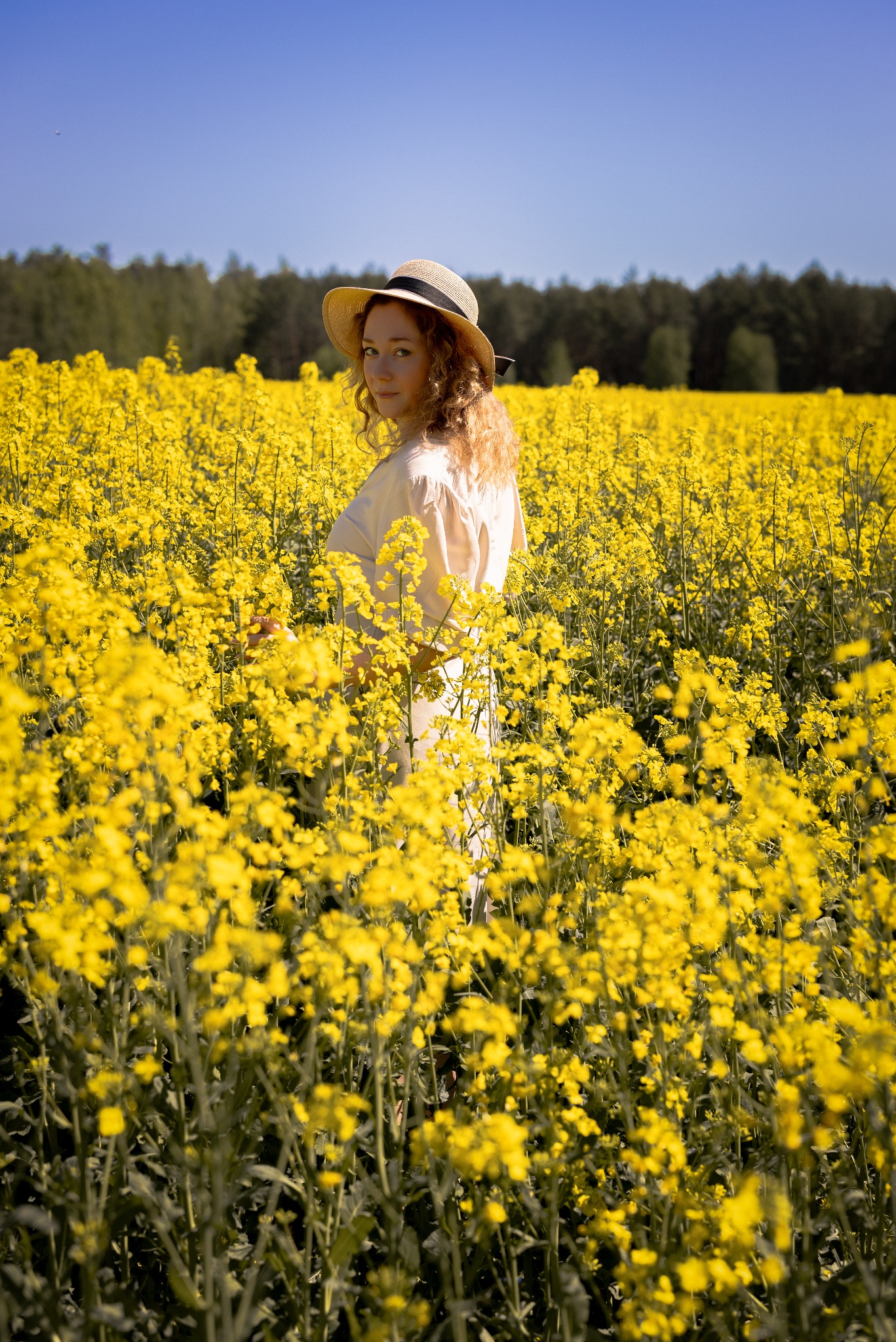 Kobieta w słomkowym kapeluszu, odwrócona tyłem, stoi w polu kwitnącego rzepaku. W tle widoczny las i błękitne niebo.