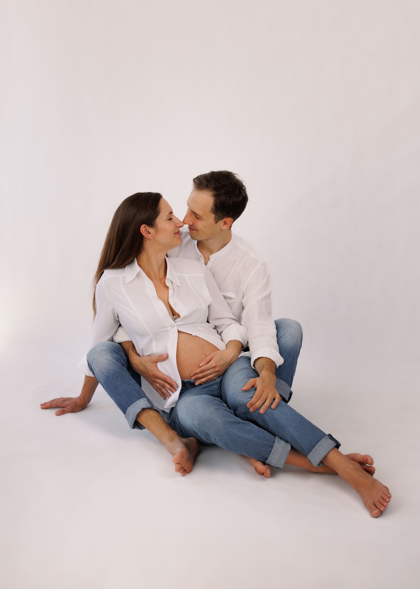 Uśmiechnięta para w białych koszulach i jeansach siedzi na podłodze, obejmując ciążowy brzuch kobiety. Delikatne oświetlenie, jasne tło. Sesja ciążowa w studio.