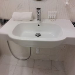 Kaczki Sprzątaczki - Biała umywalka z baterią i ręcznikiem w czystej łazience z marmurowymi płytkami. Na blacie kosmetyki hotelowe. Widok z przodu, oświetlenie górne.