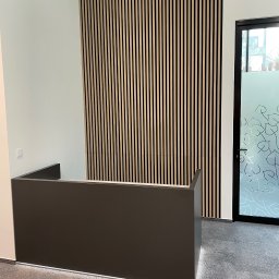 Mateusz Sulis - Nowoczesna recepcja z ciemnoszarą ladą i drewnianą ścianą z pionowych listew. W tle przeszklone drzwi z motywem roślinnym.