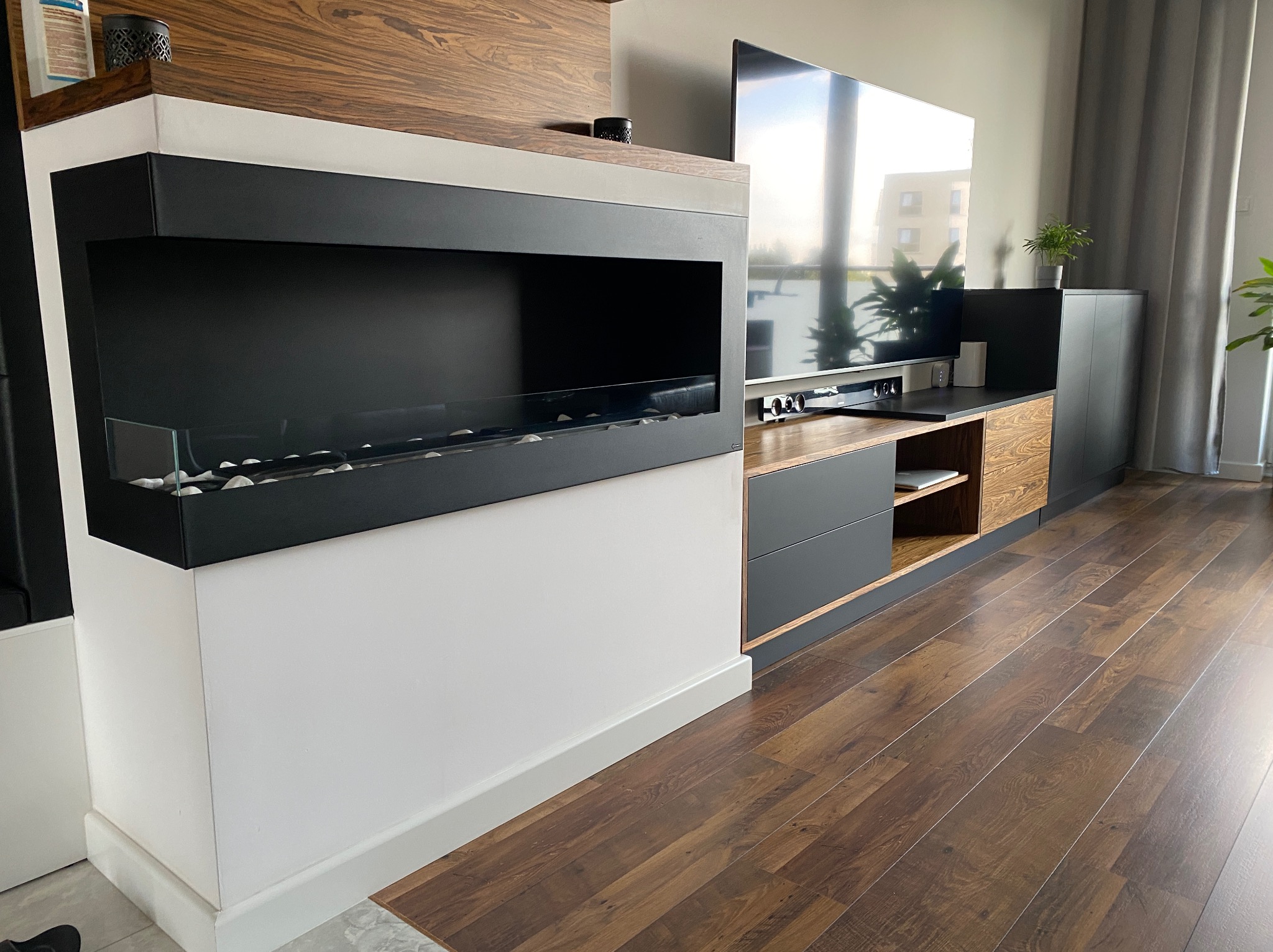 Nowoczesna zabudowa salonu z biokominkiem i szafkami w odcieniach szarości, drewna i bieli. Widoczny parkiet i okno z widokiem na budynek.