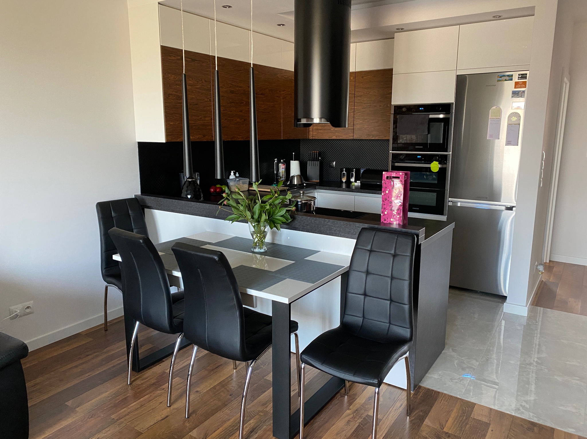 Nowoczesna kuchnia z drewnianymi akcentami, czarnym okapem i stołem z czterema krzesłami. Na blacie wazon z zielonymi gałązkami. Widoczne sprzęty AGD.