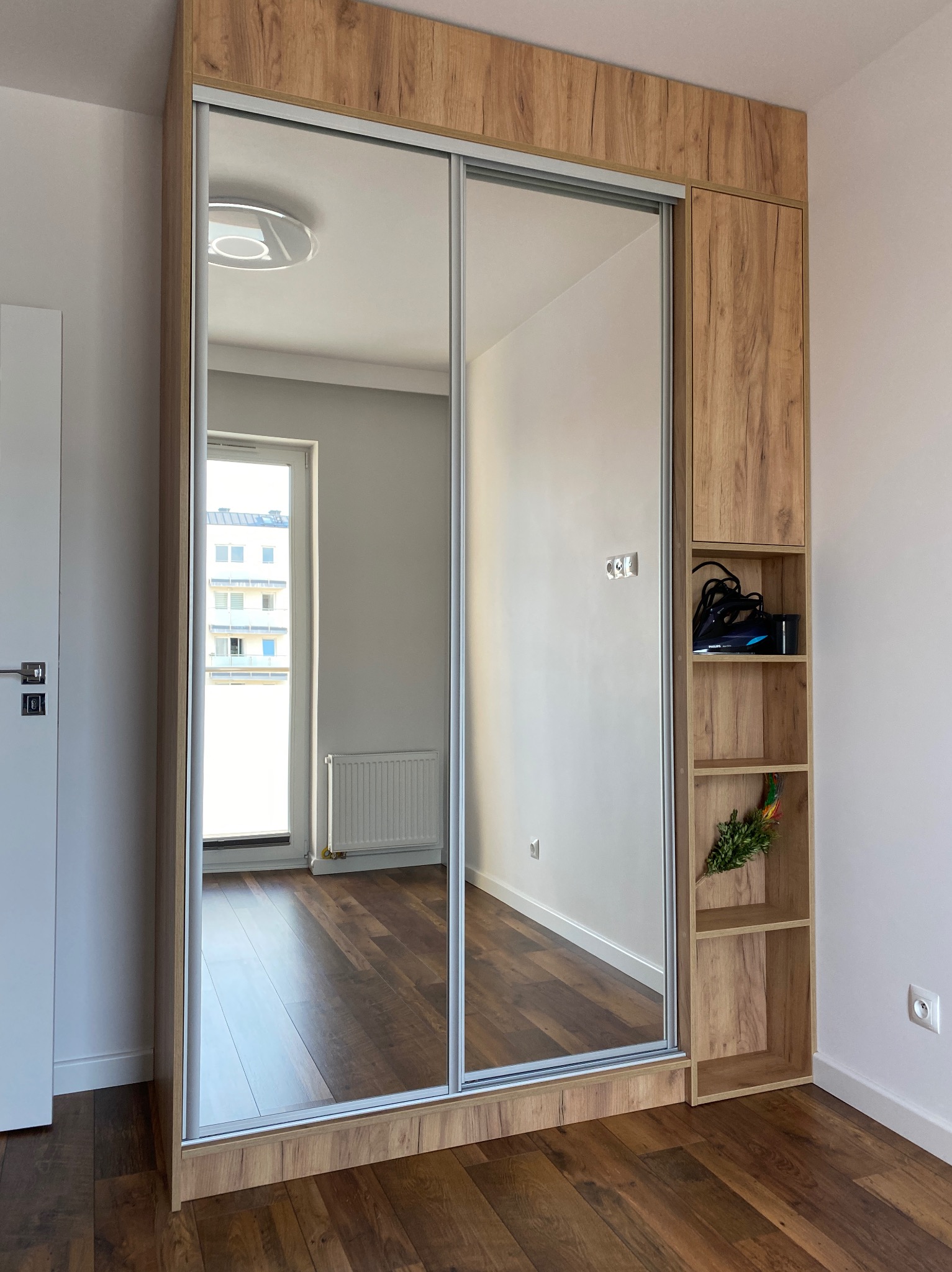 Szafa przesuwna z lustrami w drewnianej okleinie, z otwartymi półkami po prawej stronie, odbijająca wnętrze pokoju z drewnianą podłogą i oknem.