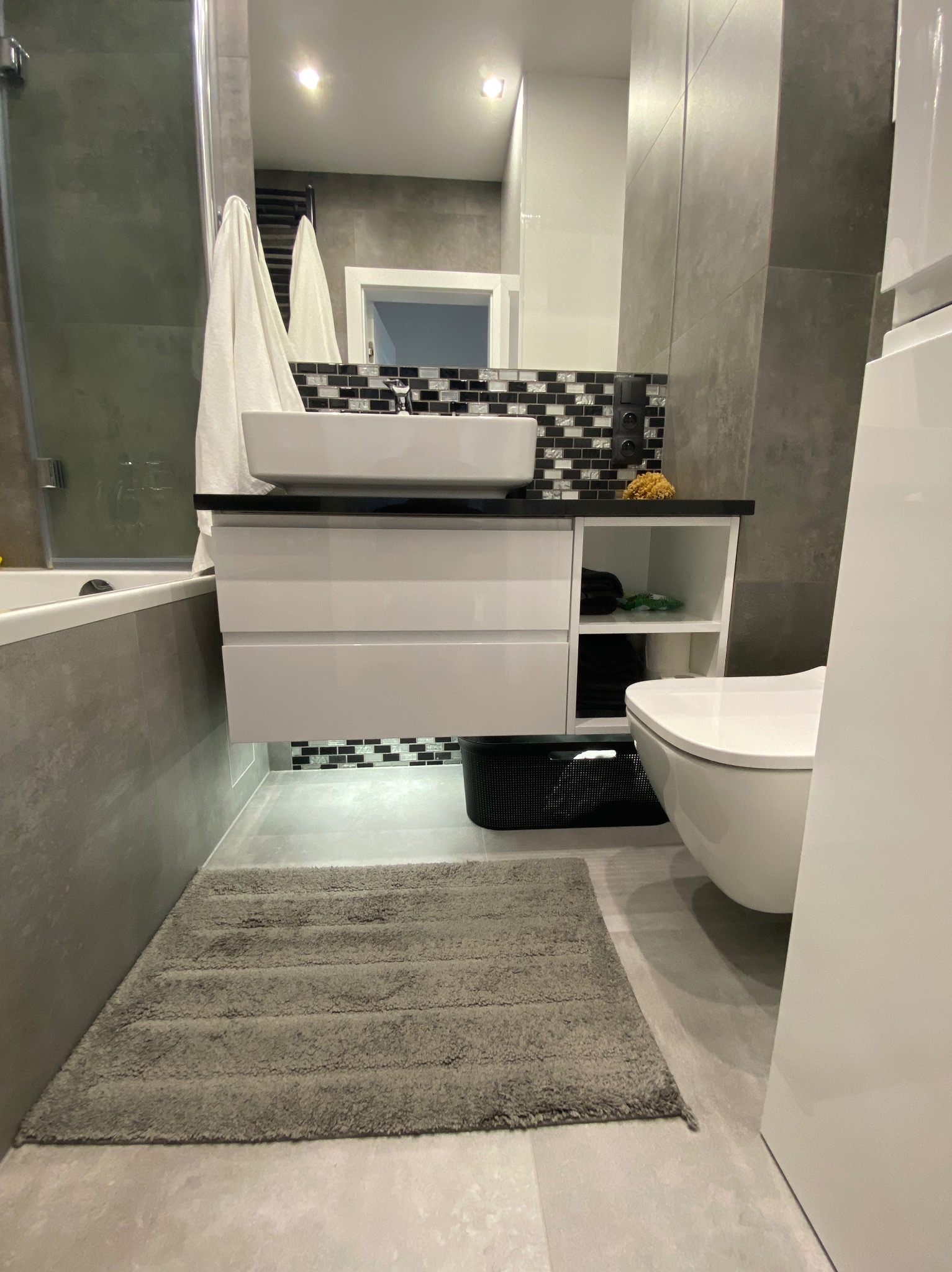 Nowoczesna łazienka z podwieszaną szafką umywalkową, mozaiką nad blatem i szarą wykładziną dywanową. Minimalistyczny design, stonowana kolorystyka.