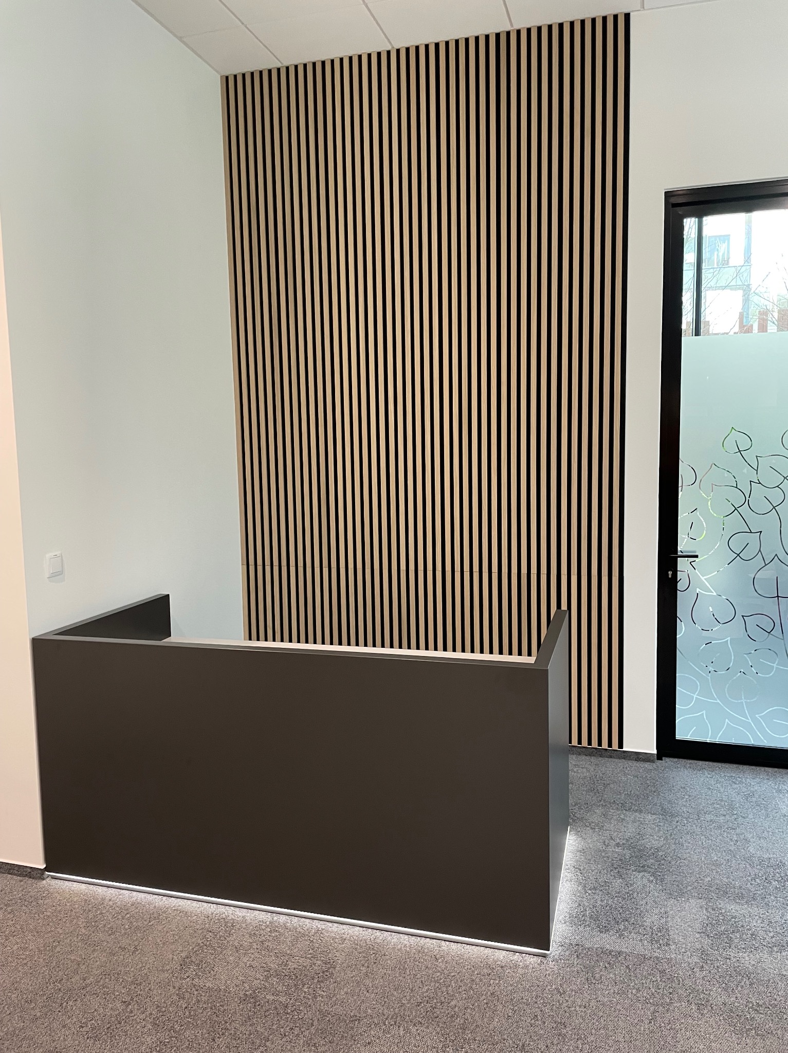Nowoczesna recepcja z ciemnoszarą ladą i drewnianą ścianą z pionowych listew. W tle przeszklone drzwi z motywem roślinnym.