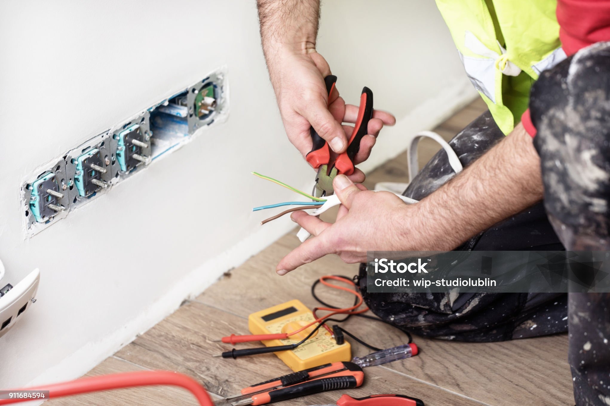 Elektryk przy pracy: montaż gniazdek w ścianie. Widoczne narzędzia, przewody i dłonie elektryka w trakcie łączenia. Ujęcie z bliska, z perspektywy podłogi.