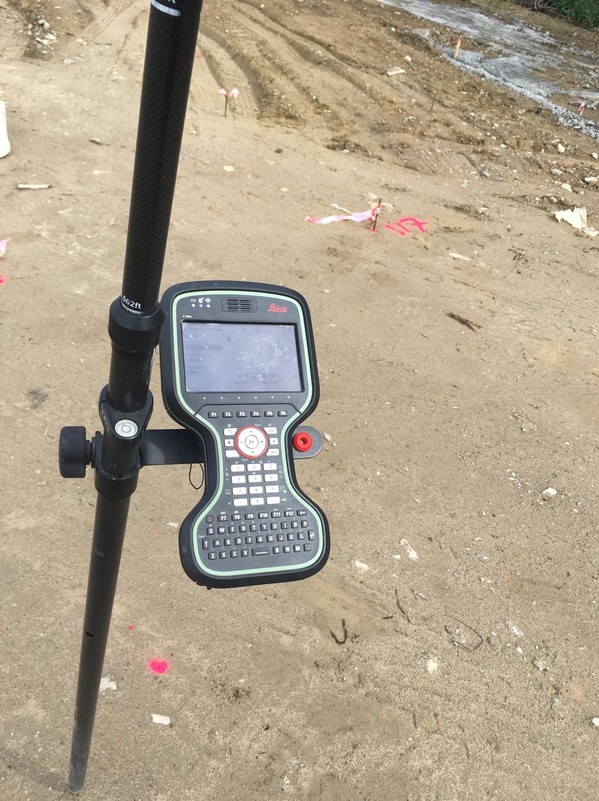 Kontroler Leica na tyczce geodezyjnej, teren budowy z oznaczeniami. Sprzęt pomiarowy w trakcie pracy, widoczne oznaczenia w terenie.