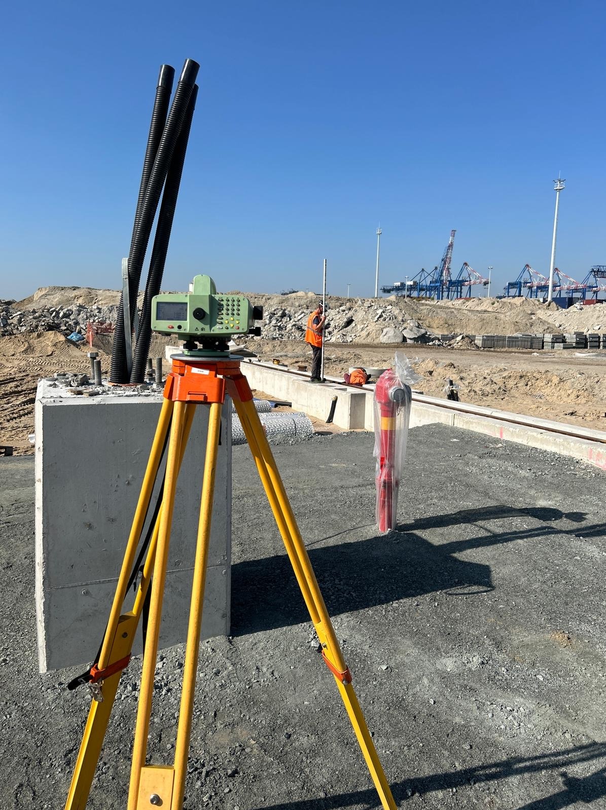 Prace pomiarowe na budowie portu: niwelator na statywie, pracownik z łatą geodezyjną, w tle dźwigi portowe i błękitne niebo. Precyzyjne wyznaczanie poziomu.