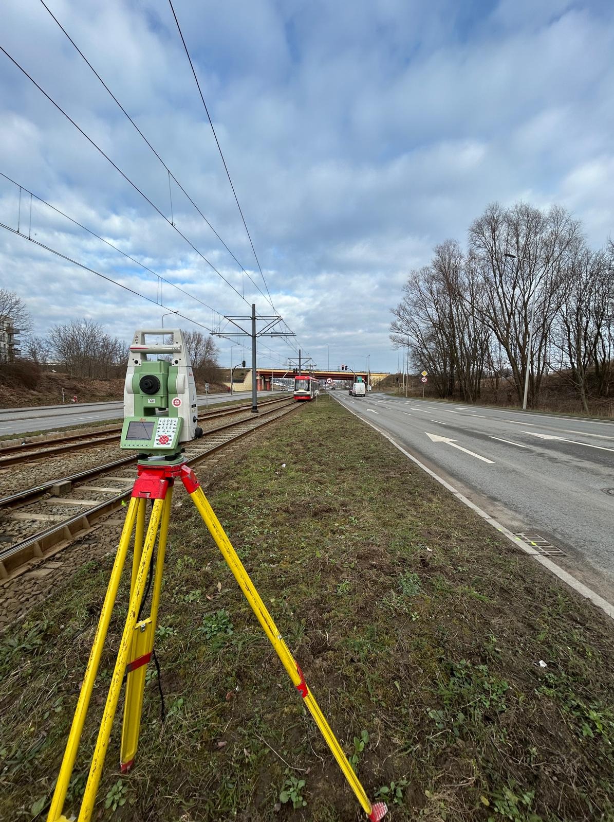 Aparat pomiarowy na żółtym statywie przy torach tramwajowych i drodze, w tle wiadukt i pochmurne niebo. Sprzęt ustawiony do pomiarów.