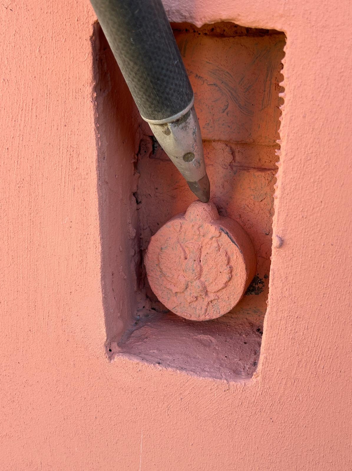 Różowa ściana z wnęką, w której znajduje się okrągły element z wytłoczonym wzorem, dotykany przez metalową końcówkę sondy geodezyjnej. Detal pomiaru geodezyjnego.