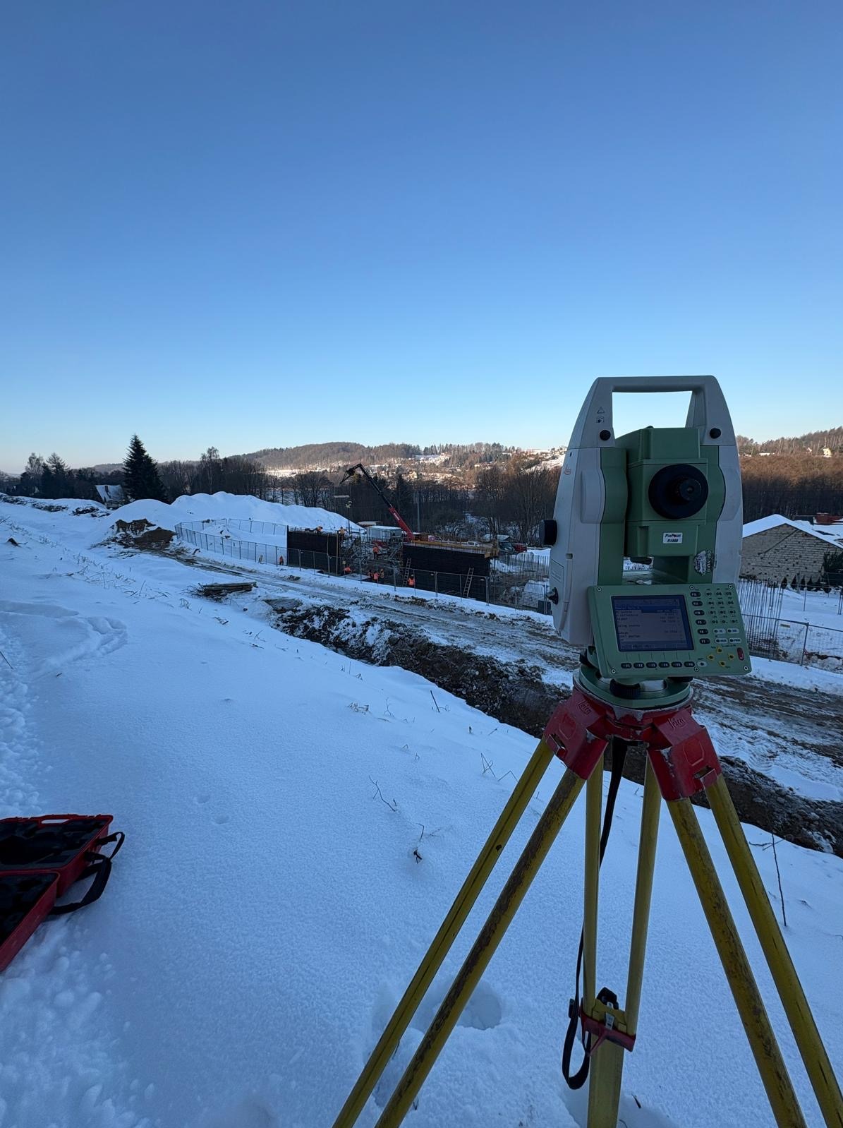 Praca w terenie zimą w Banino: Teodolit na statywie, w tle wykop i prace budowlane. Błękitne niebo, śnieg. Precyzyjne pomiary geodezyjne.