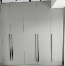 M.T.Meble Na Wymiar Mariusz Turkowski - Szara szafa wnękowa z czarnymi, pionowymi uchwytami, oświetlona reflektorami sufitowymi. Minimalistyczny design, jasna podłoga.