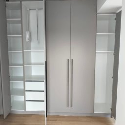 M.T.Meble Na Wymiar Mariusz Turkowski - Jasnoszara szafa wnękowa z czarnymi uchwytami, częściowo otwarta, ukazująca białe półki i szuflady. Oświetlenie punktowe na suficie. Parkiet na podłodze.