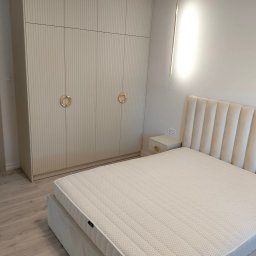 M.T.Meble Na Wymiar Mariusz Turkowski - Jasna szafa wnękowa z ryflowanymi frontami i złotymi uchwytami w sypialni z łóżkiem i lampą. Minimalistyczny design, ciepłe oświetlenie.