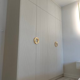 M.T.Meble Na Wymiar Mariusz Turkowski - Biała szafa wnękowa z ryflowanymi frontami i złotymi, okrągłymi uchwytami, dopasowana do wnętrza sypialni. Minimalistyczny design, jasne drewno na podłodze.