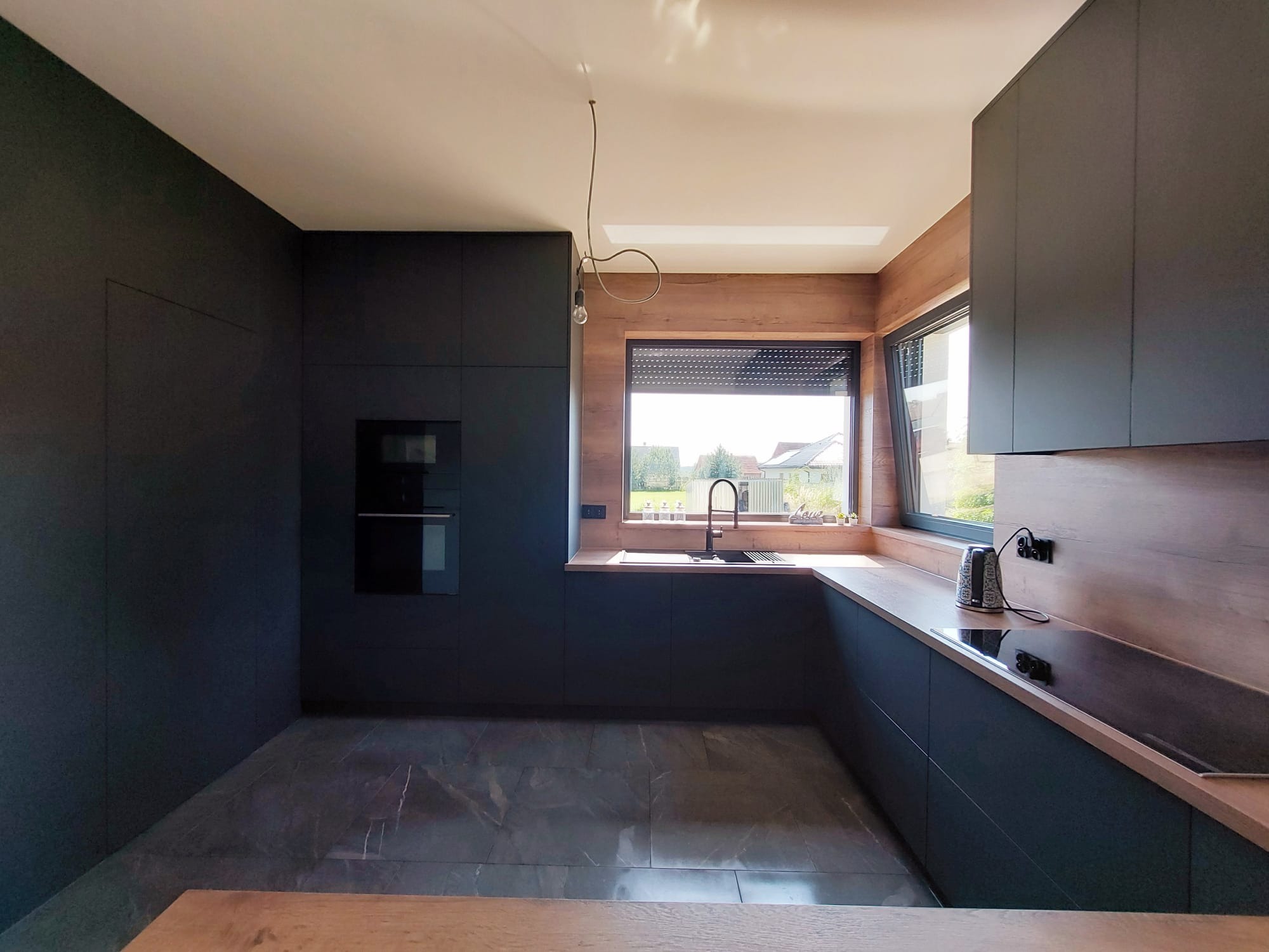 Nowoczesna kuchnia z ciemnymi, matowymi frontami i drewnianymi akcentami. Widoczne okno z roletami i minimalistyczna lampa wisząca. Marmurowa podłoga dodaje elegancji.