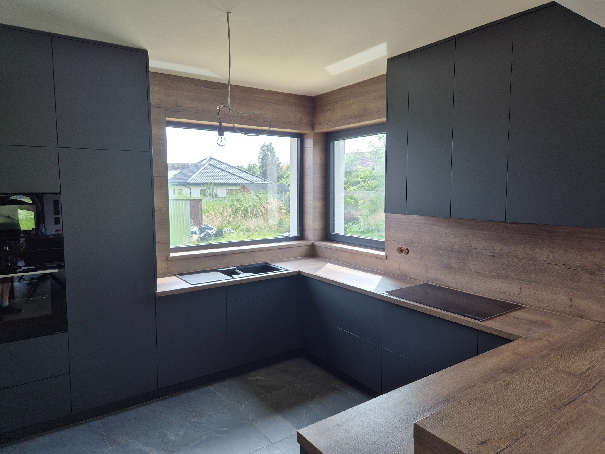Nowoczesna kuchnia w kształcie litery U z ciemnymi frontami i drewnianym blatem, oświetlona naturalnym światłem w Twardogórze. Widok na ogród przez okno.