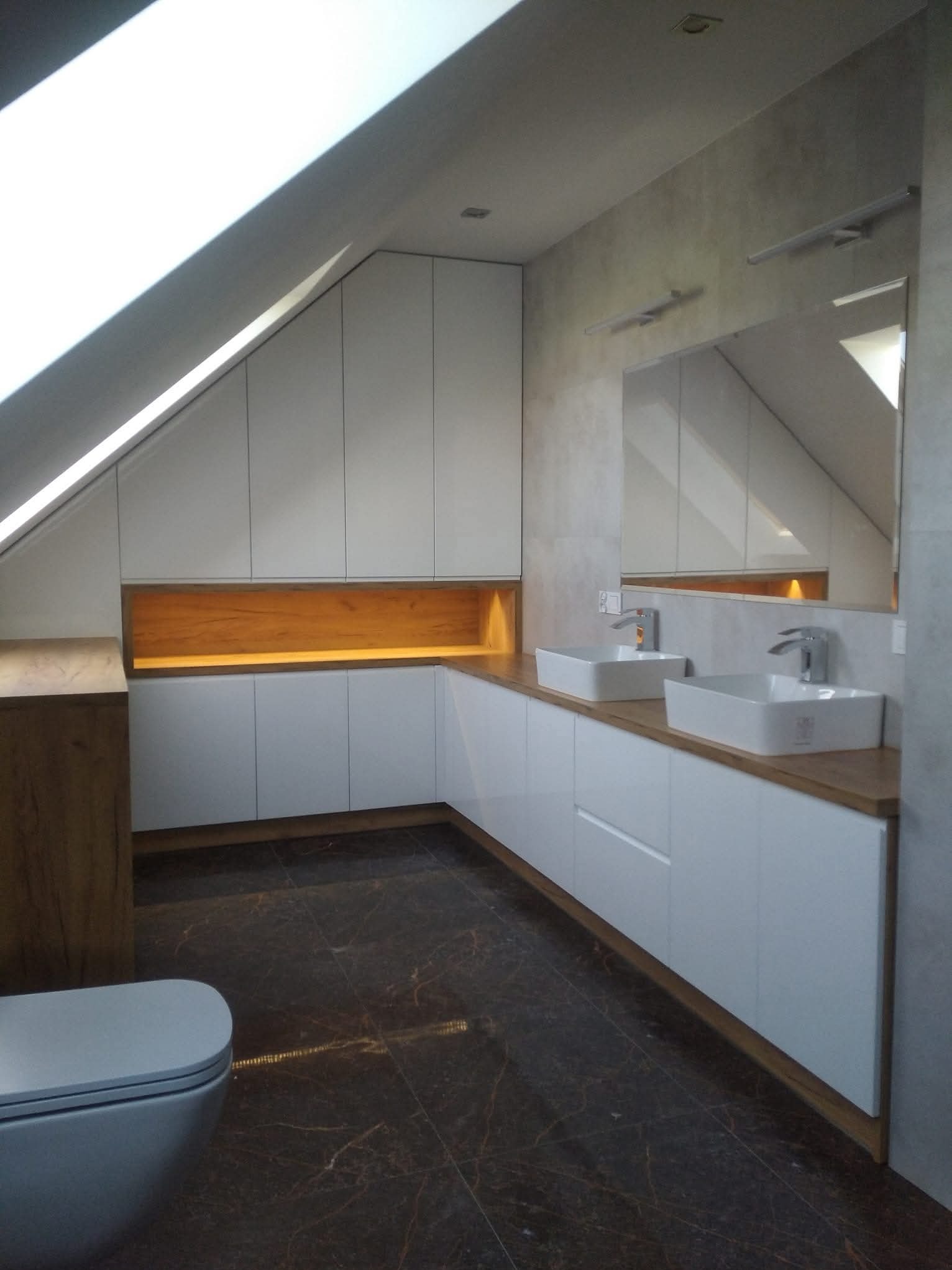 Białe szafki łazienkowe pod skosem dachu z drewnianym blatem i oświetleniem LED, dwie umywalki nablatowe, duże lustro, ciemna marmurowa podłoga. Nowoczesna aranżacja.