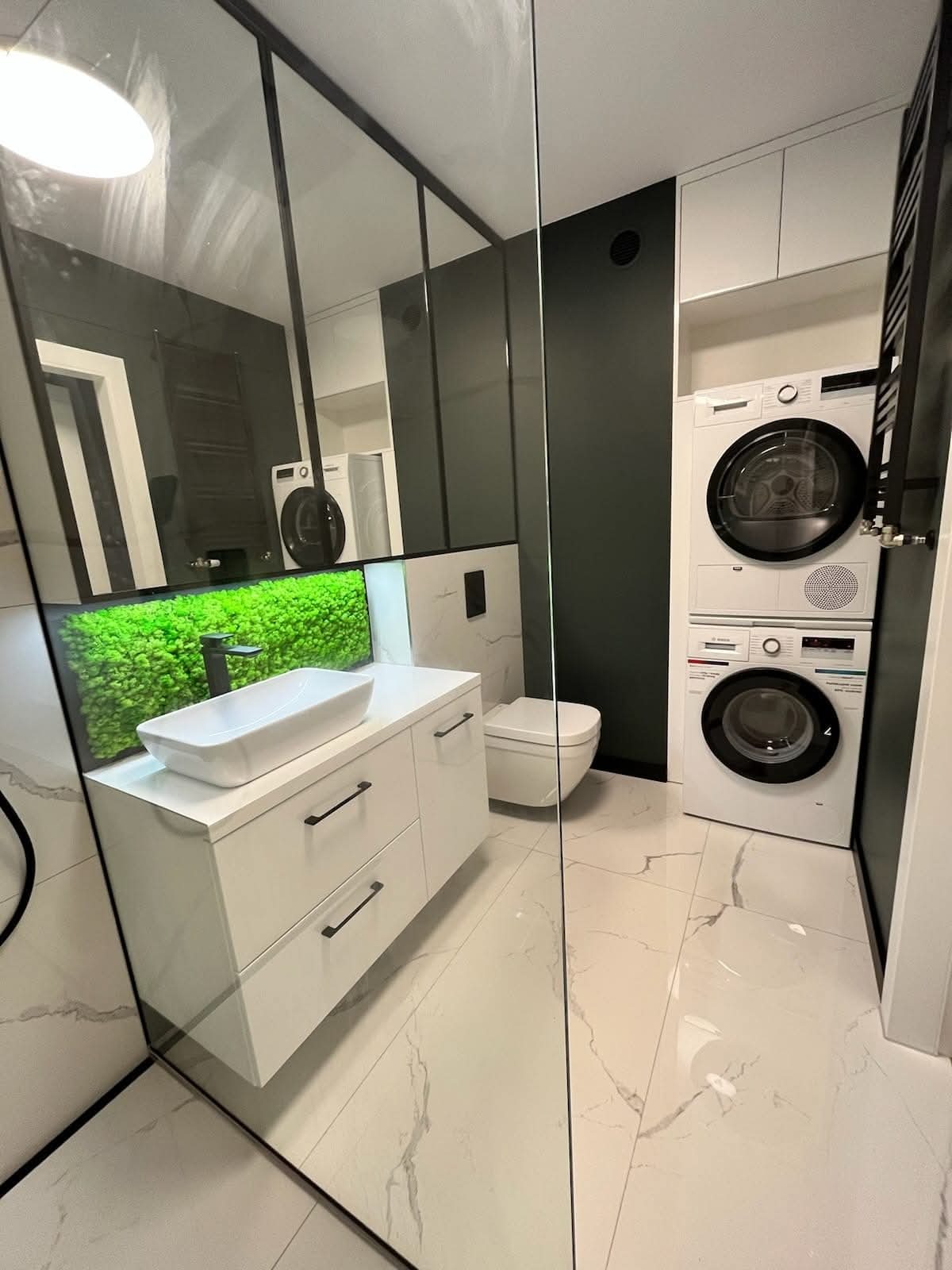 Nowoczesna łazienka z białą szafką pod umywalkę, zieloną ścianą z mchu, toaletą i pralko-suszarką w zabudowie. Marmurowe płytki podłogowe.