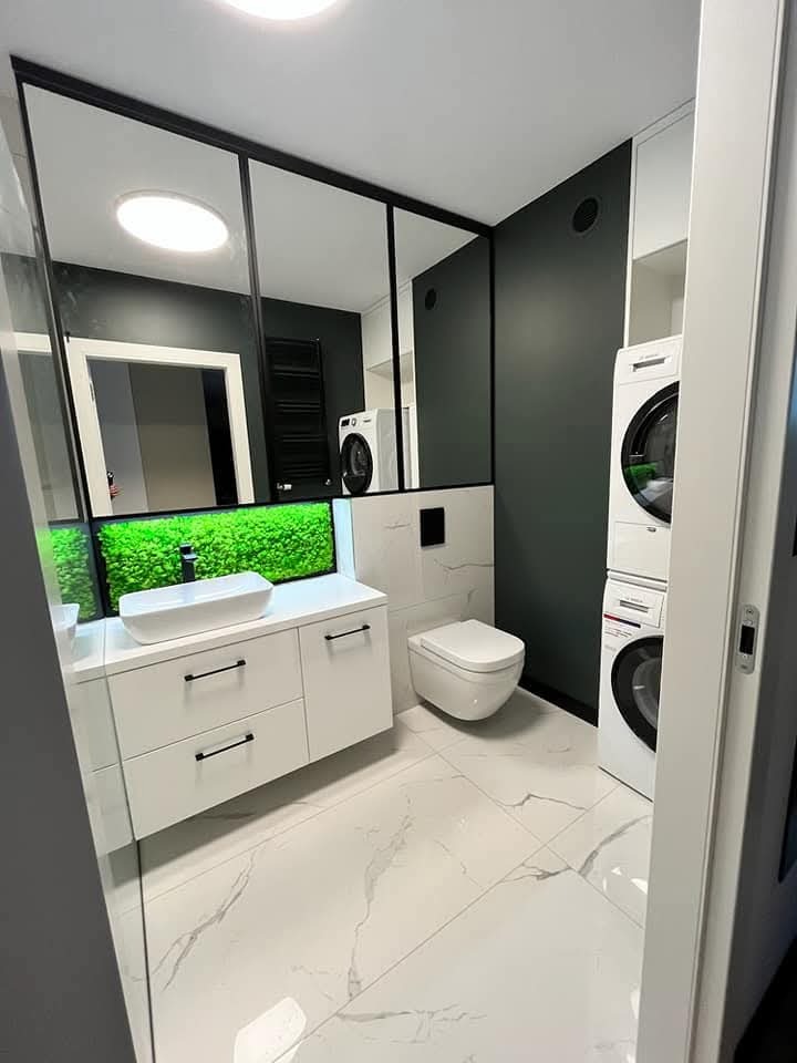 Nowoczesna łazienka z białą szafką pod umywalkę, toaletą i pralko-suszarką w zabudowie. Lustro z czarną ramą i podświetleniem LED. Marmurowe płytki podłogowe.