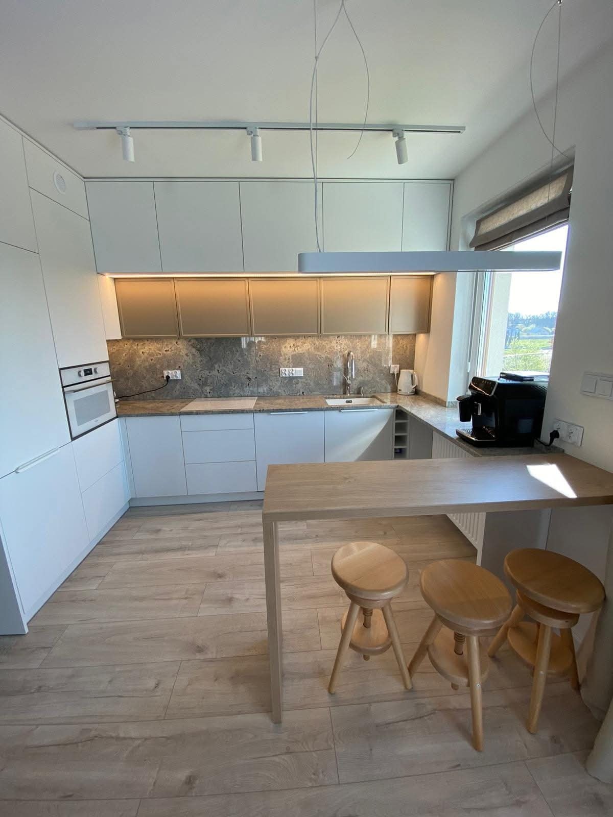 Jasna kuchnia z drewnianym blatem i hokerami. Minimalistyczny design, szare blaty, oświetlenie punktowe i wiszące lampy. Ekspres do kawy na blacie.