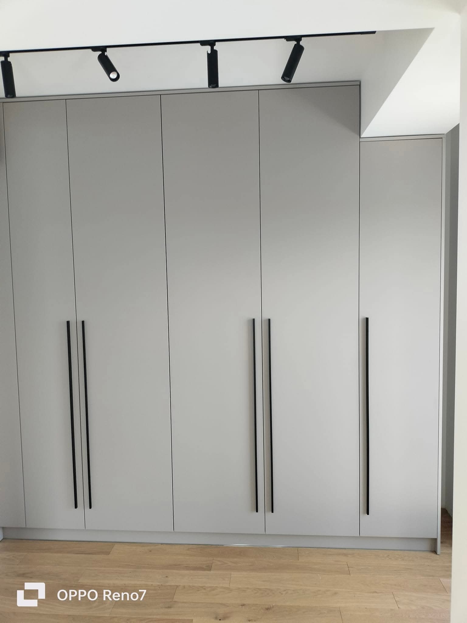 Szara szafa wnękowa z czarnymi, pionowymi uchwytami, oświetlona reflektorami sufitowymi. Minimalistyczny design, jasna podłoga.