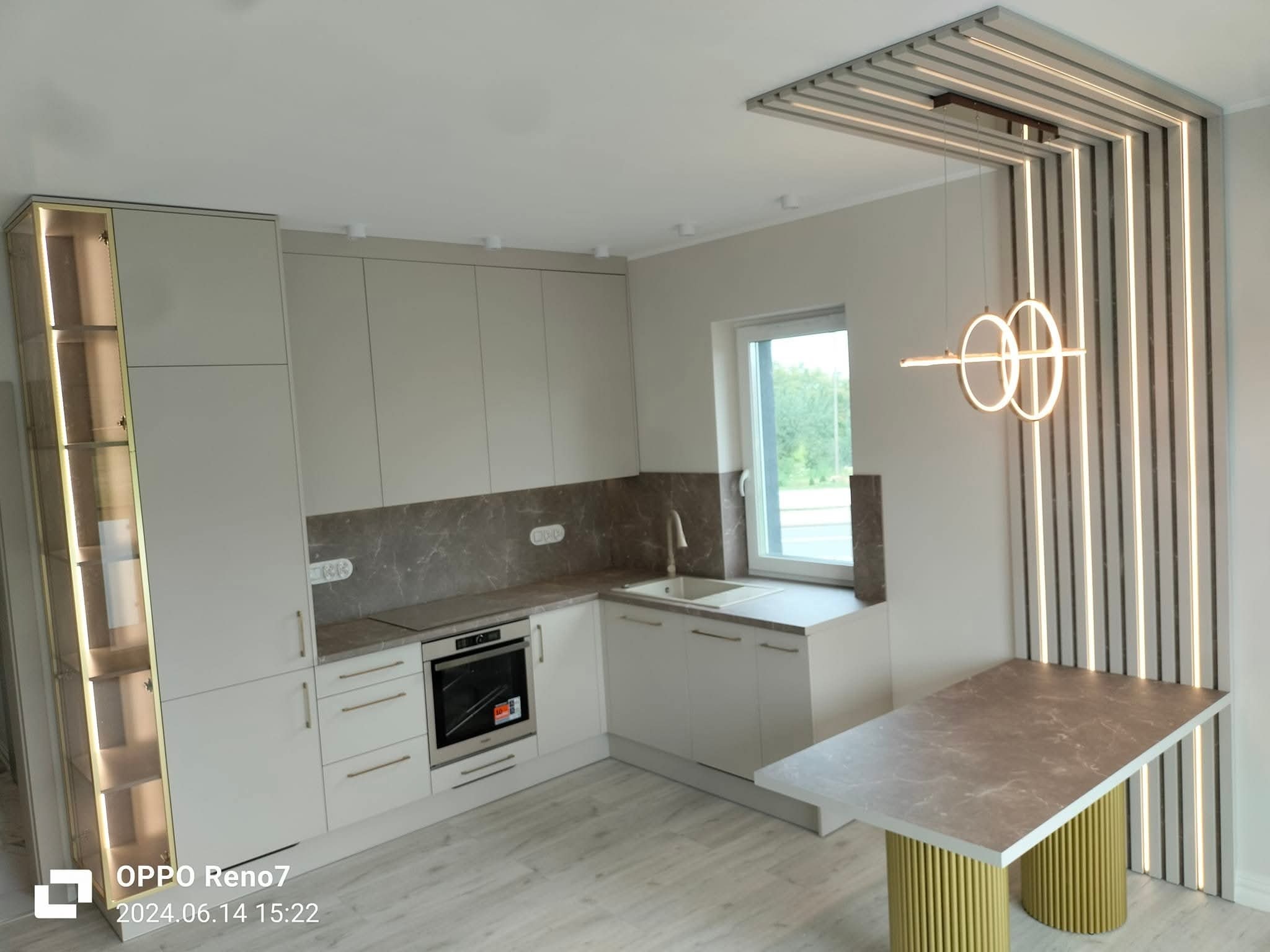 Jasna kuchnia z marmurowym blatem i złotymi akcentami, oświetlona lampą w kształcie okręgów. Widoczny fragment stołu z blatem pasującym do blatu kuchennego.