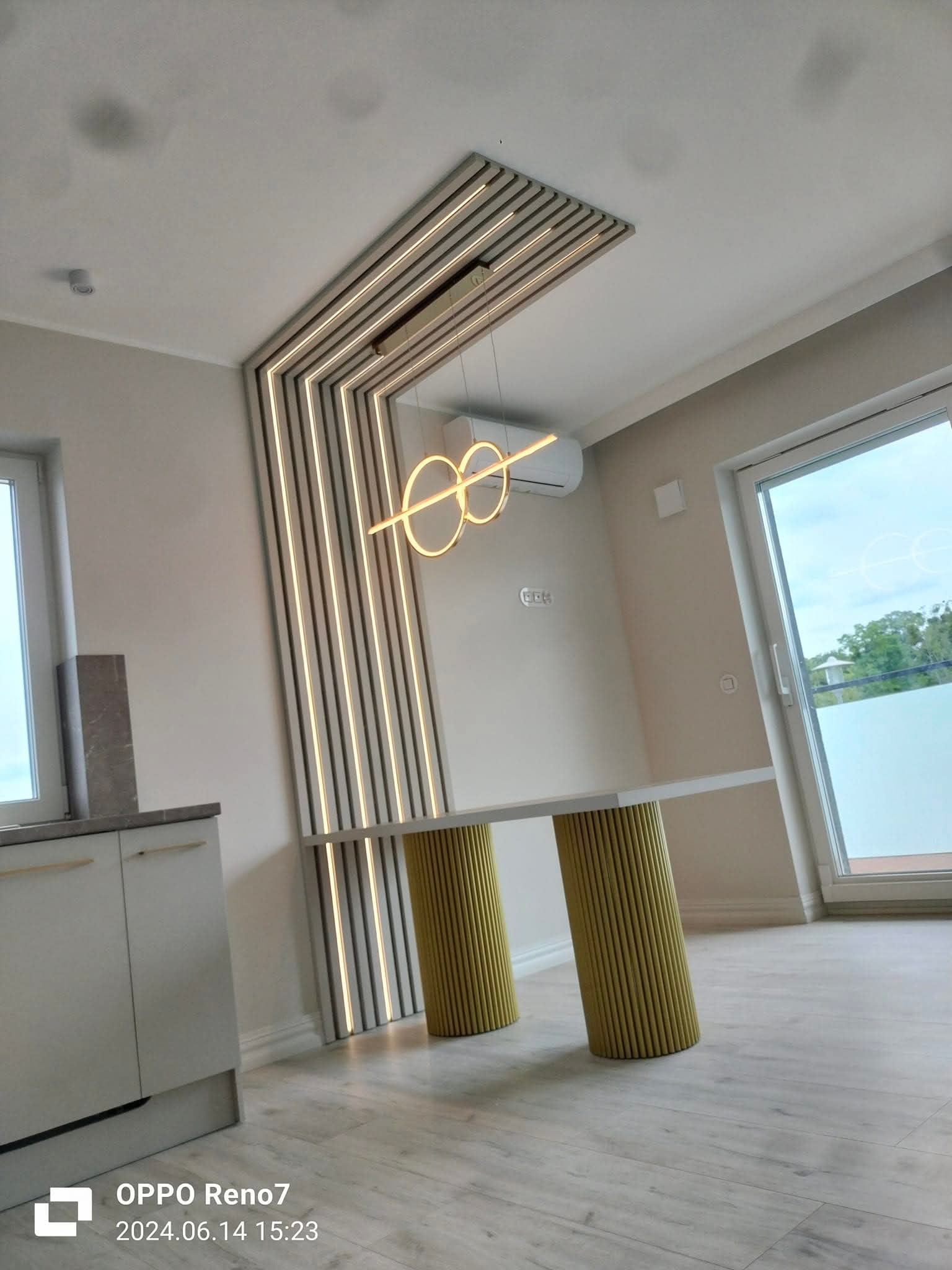 Nowoczesna aranżacja wnętrza z elementami dekoracyjnymi: podświetlane lamele ścienne, designerska lampa i stół z żółtymi nogami. Widok na okno i fragment zabudowy kuchennej.