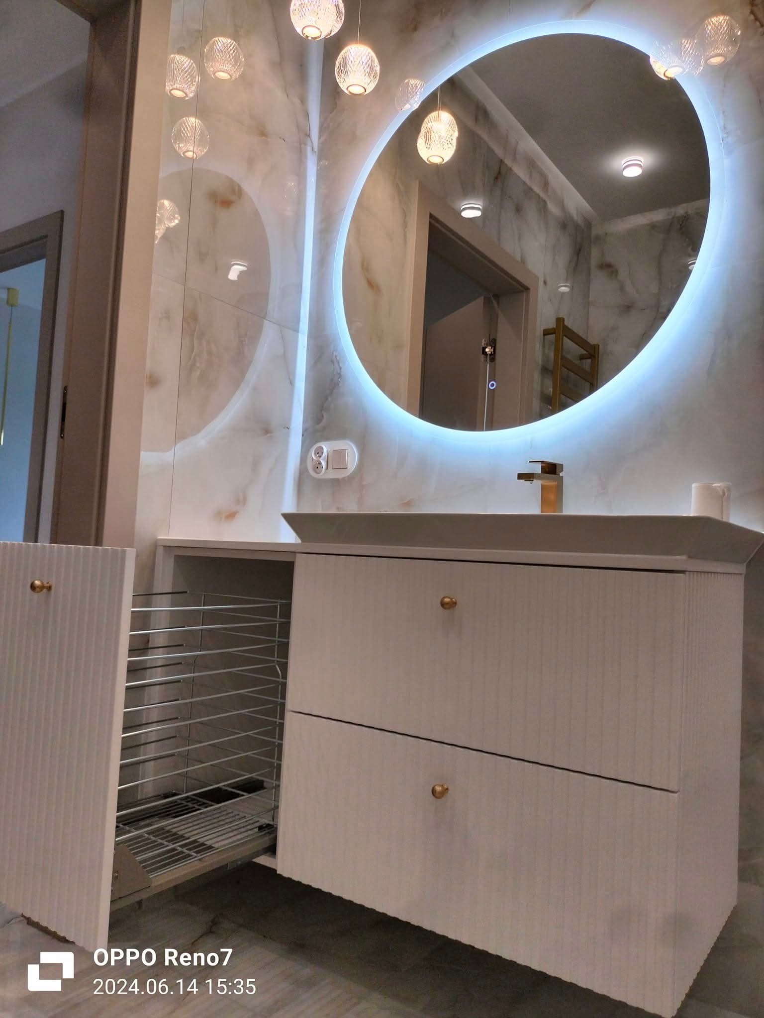 Biała szafka łazienkowa z wysuwanym koszem i owalnym lustrem z podświetleniem LED, marmurowe ściany, złota bateria. Nowoczesny design.