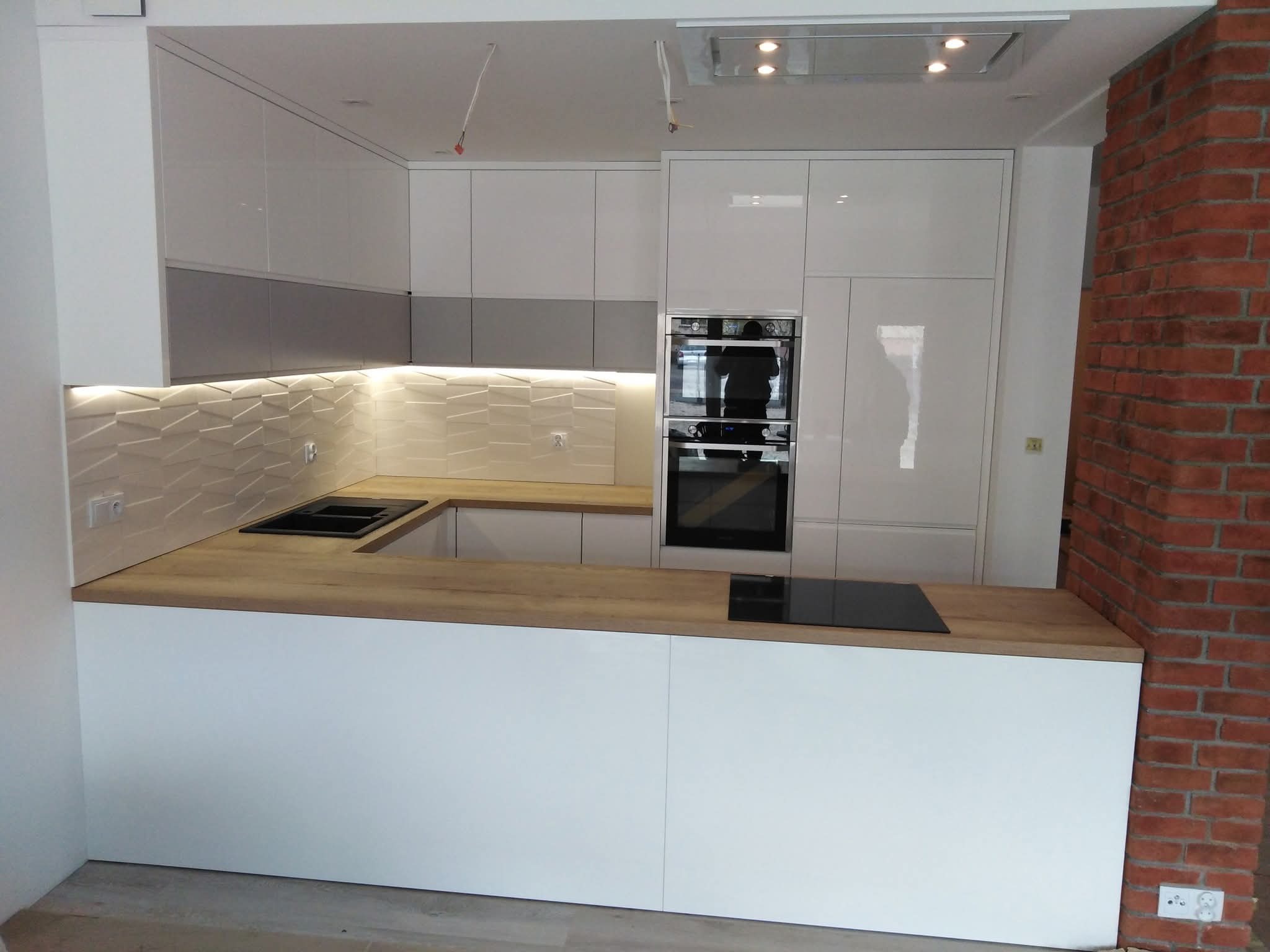 Biała kuchnia z drewnianym blatem i ceglaną ścianą. Nowoczesne szafki z połyskiem i oświetleniem LED. Minimalistyczny design z płytą indukcyjną.
