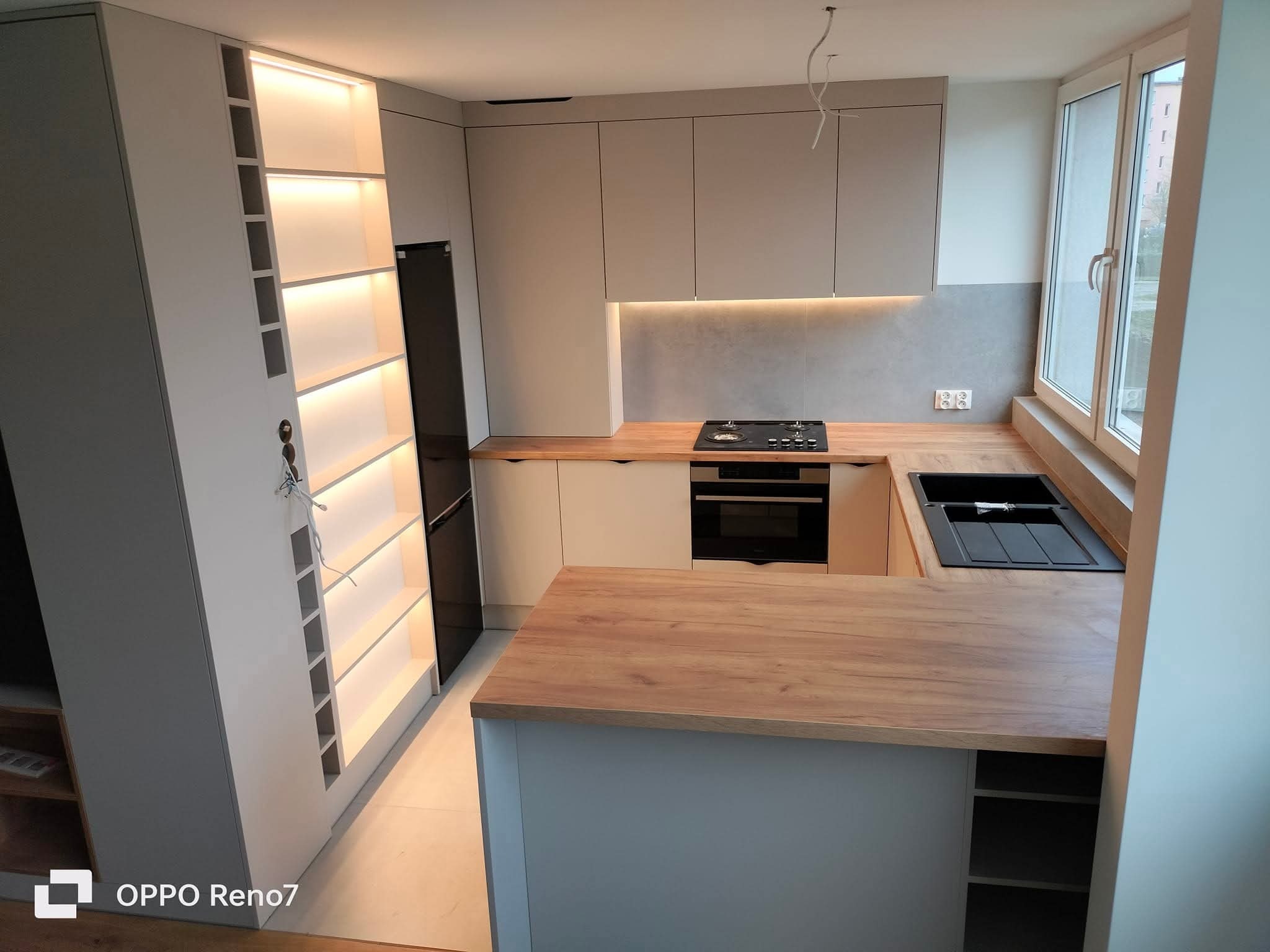 Nowoczesna kuchnia z wyspą i oświetleniem LED w półkach. Jasne fronty szafek, drewniany blat i czarny zlew. Widoczne gniazdka elektryczne i fragment okna.