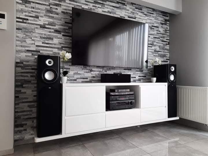 Biała szafka RTV z czarnymi głośnikami na tle ściany z szarej cegły, pod telewizorem. Minimalistyczny design, nowoczesne wnętrze z kwiatami w doniczkach.