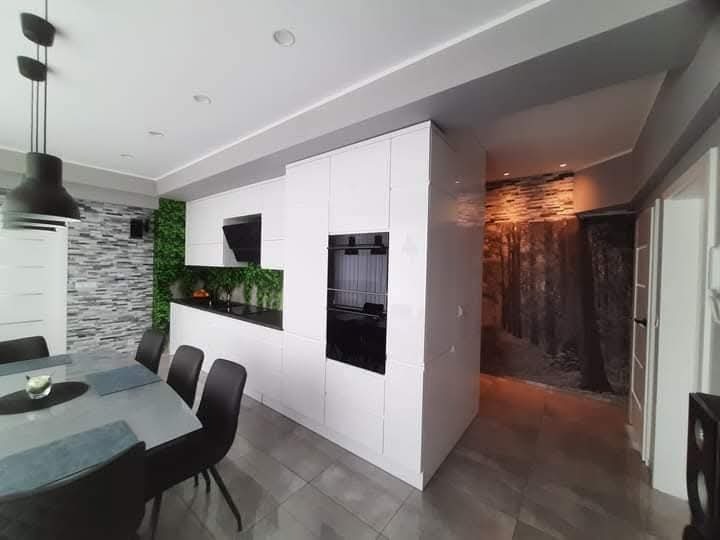 Nowoczesna, biała kuchnia z czarnymi akcentami, widok z jadalni. Na ścianie dekoracyjny panel z zielenią i kamieniem, w tle fototapeta z motywem leśnym.