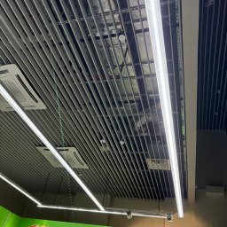 Szo Ne Bud - Sufit rastrowy z oświetleniem LED i klimatyzacją w przestrzeni komercyjnej. Widoczne elementy instalacji nad sufitem. Ujęcie z dołu, pod kątem.