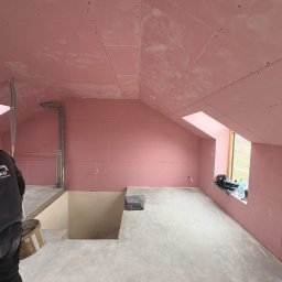 SZO-NE-BUD OLEH PYLYPCHUK - Poddasze w trakcie wykańczania: ściany i skosy obłożone różową płytą, widoczne łączenia i otwory okienne. Na podłodze resztki materiałów budowlanych.
