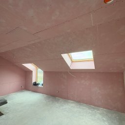 SZO-NE-BUD OLEH PYLYPCHUK - Pokój na poddaszu z oknami dachowymi, wykończony płytami kartonowo-gipsowymi w kolorze różowym, przygotowany do dalszych prac wykończeniowych. Widoczny surowy beton na podłodze.