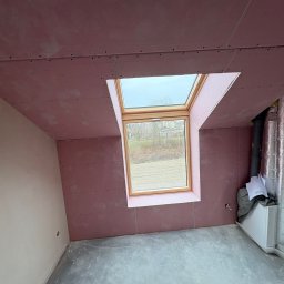 SZO-NE-BUD OLEH PYLYPCHUK - Poddasze z oknem dachowym w trakcie wykańczania: różowe płyty kartonowo-gipsowe na ścianach i suficie, widoczne łączenia, surowa podłoga, instalacja wentylacyjna.